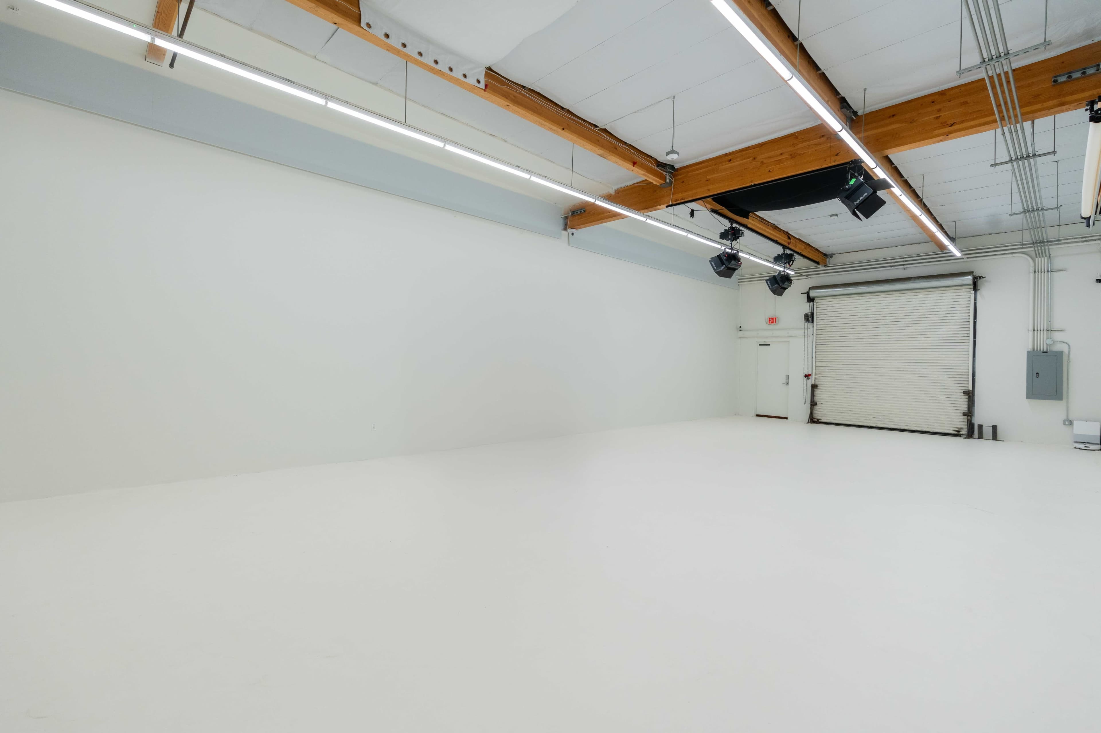 The image shows an empty studio space with white walls, a white floor, wooden beams, and a closed rolling door.