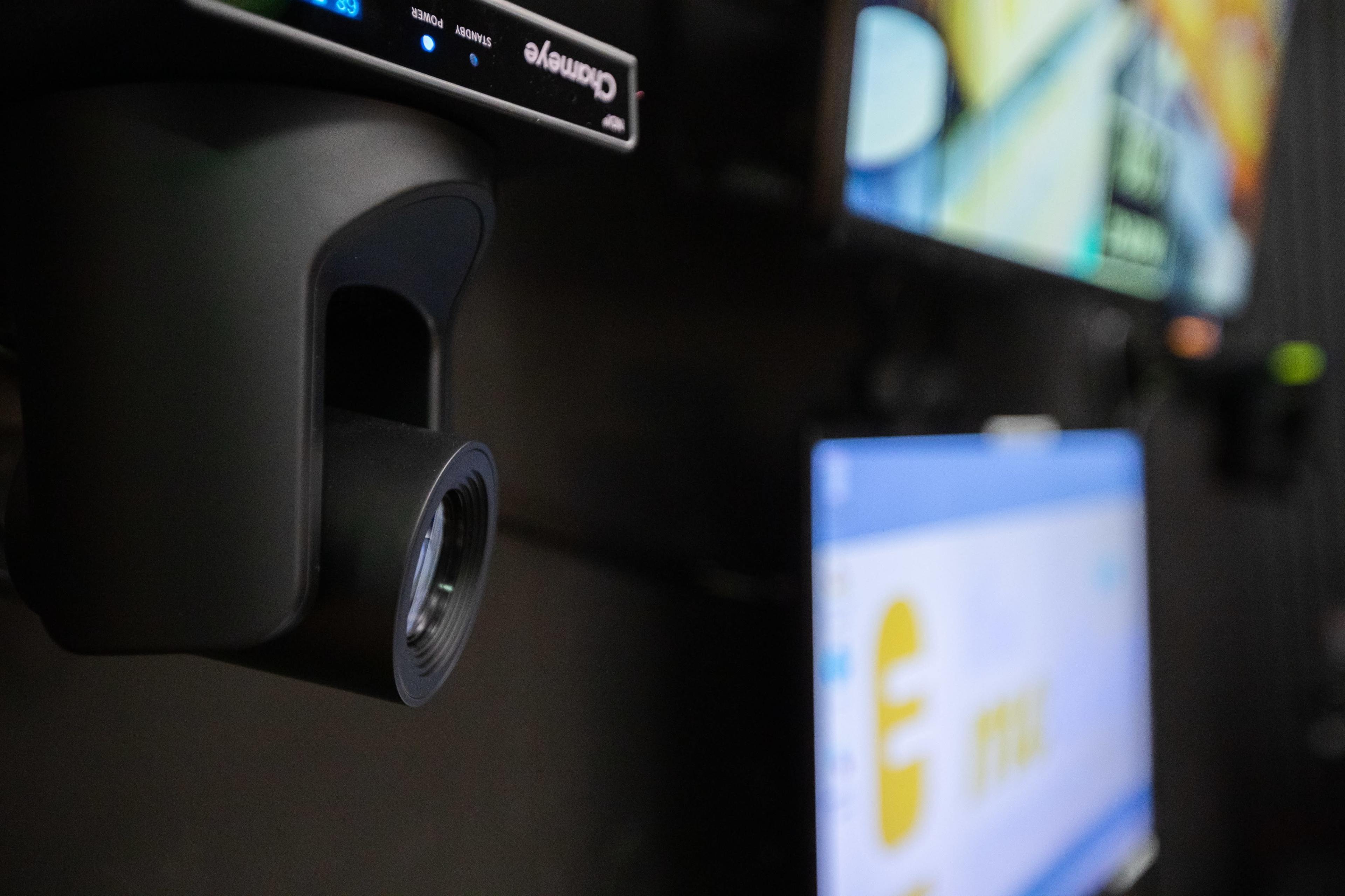 A black camera is mounted on a wall beside multiple monitors displaying various graphics and information.