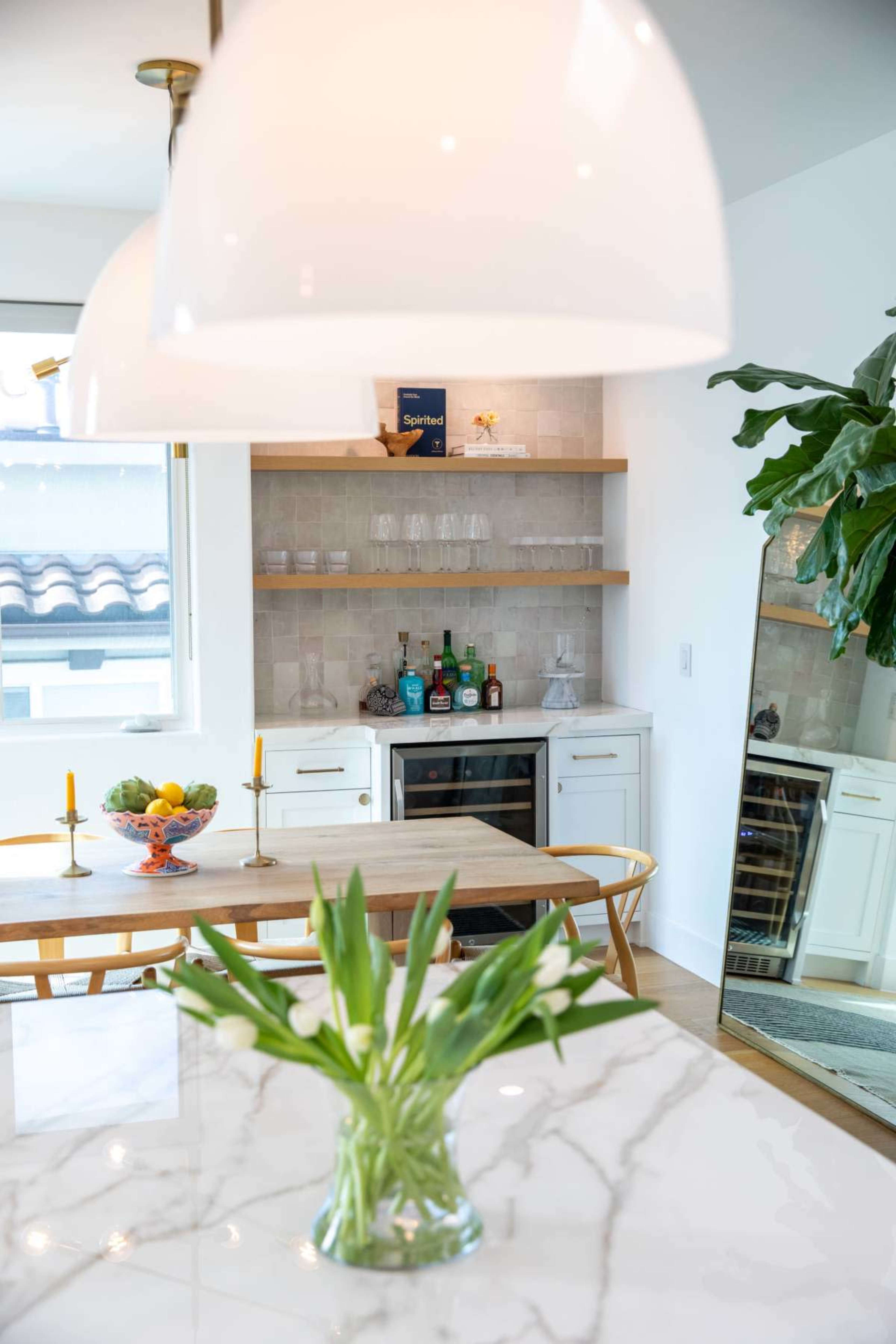 A modern kitchen features a dining table with fruit, a bar area with assorted drinks, and a large mirror reflecting the space.