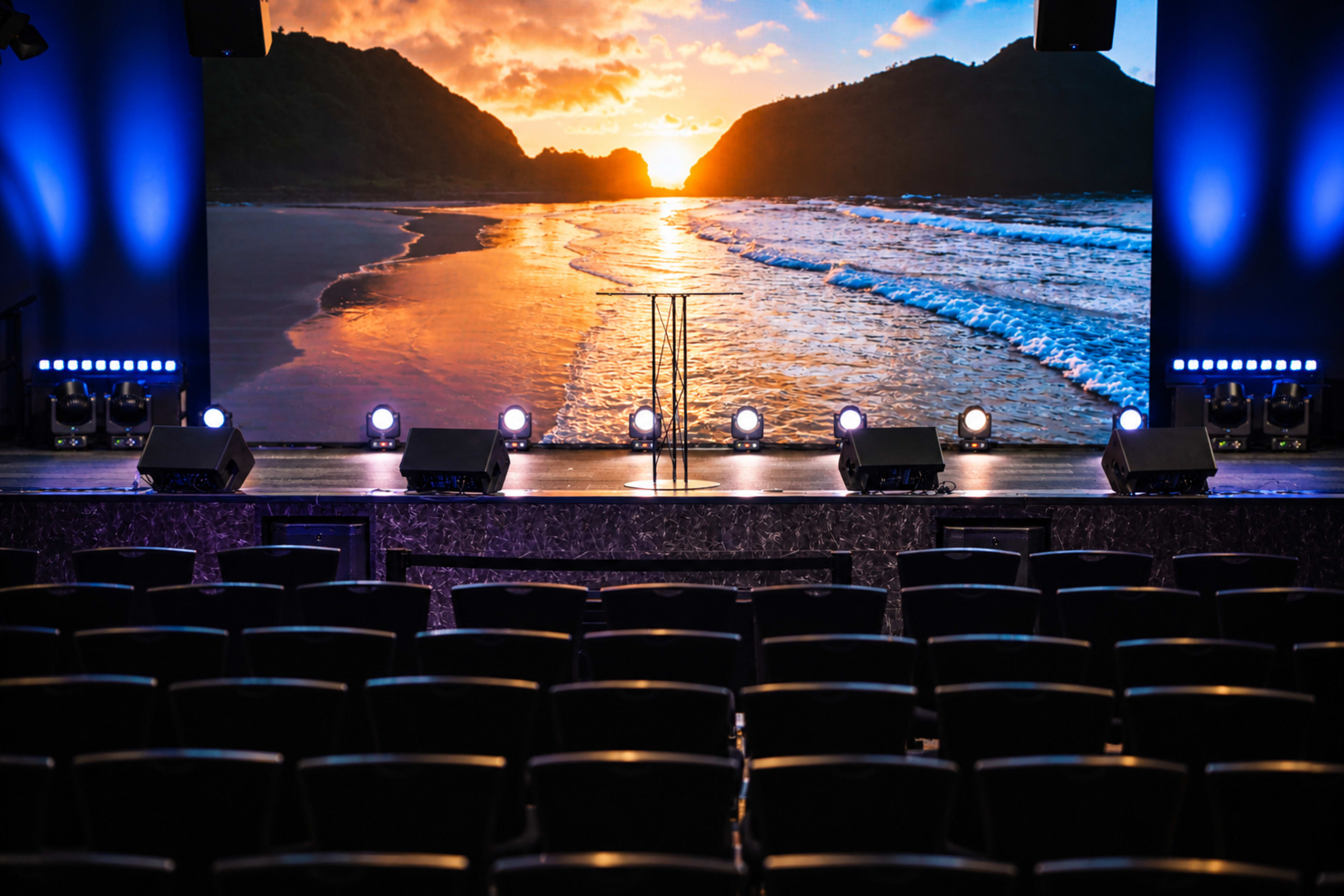The image shows an auditorium setting with rows of empty black chairs facing a stage that features a large backdrop of a sunset over a beach, accompanied by stage lights.