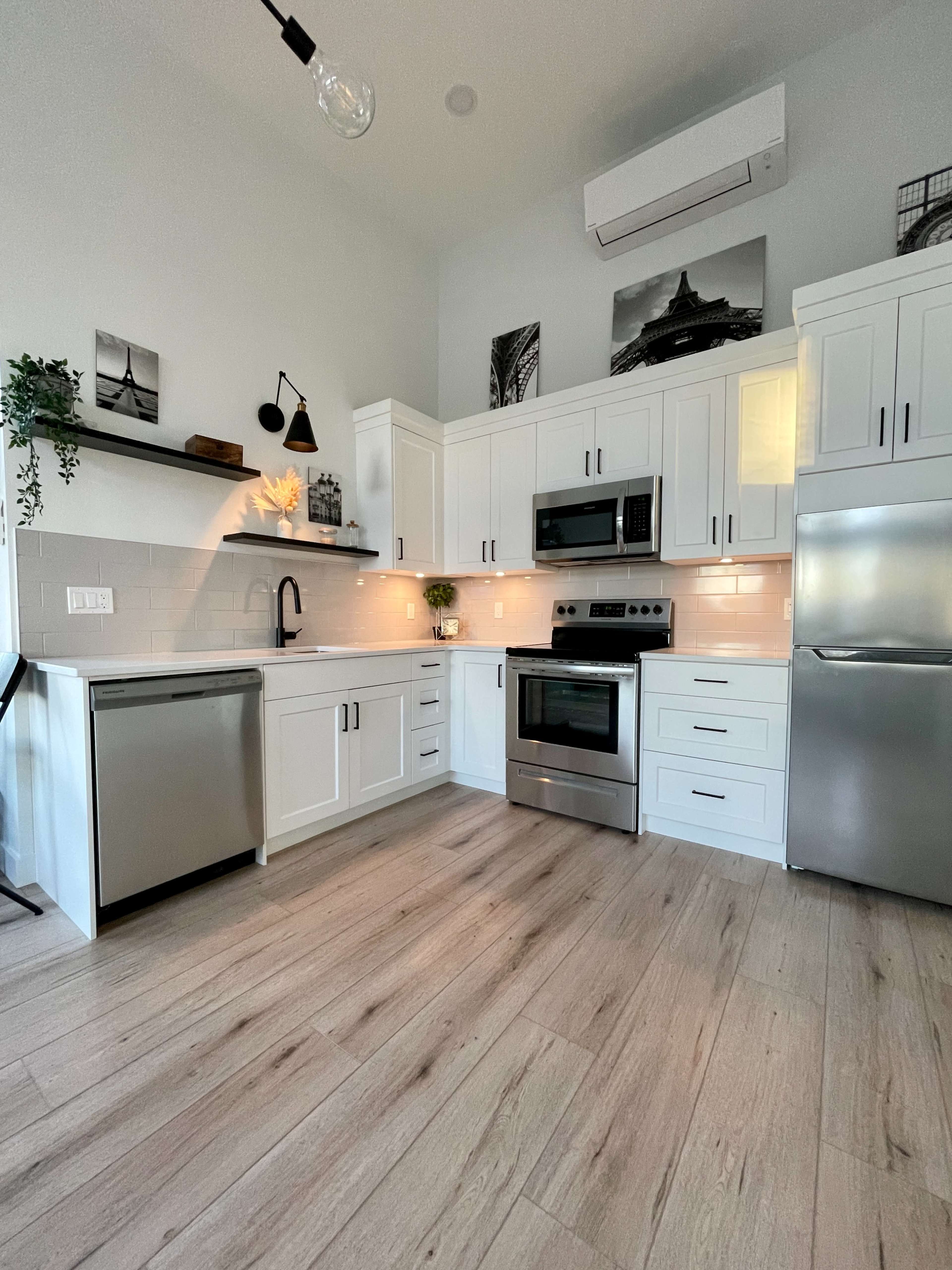 The kitchen features white cabinets, stainless steel appliances, and a light wood floor, with minimalistic decor on the walls.