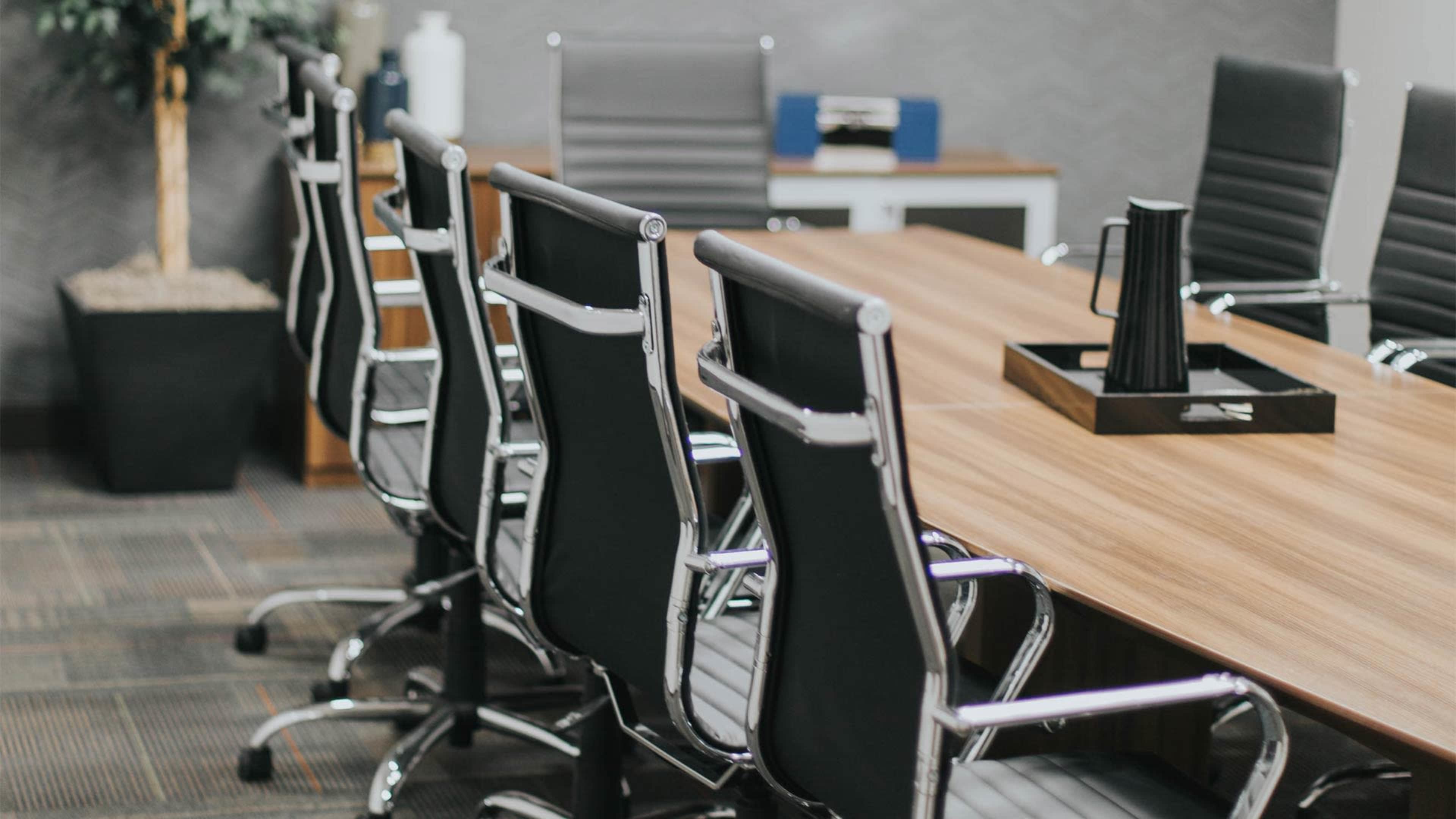 A modern conference room features a long wooden table surrounded by black office chairs and a potted plant in the corner.