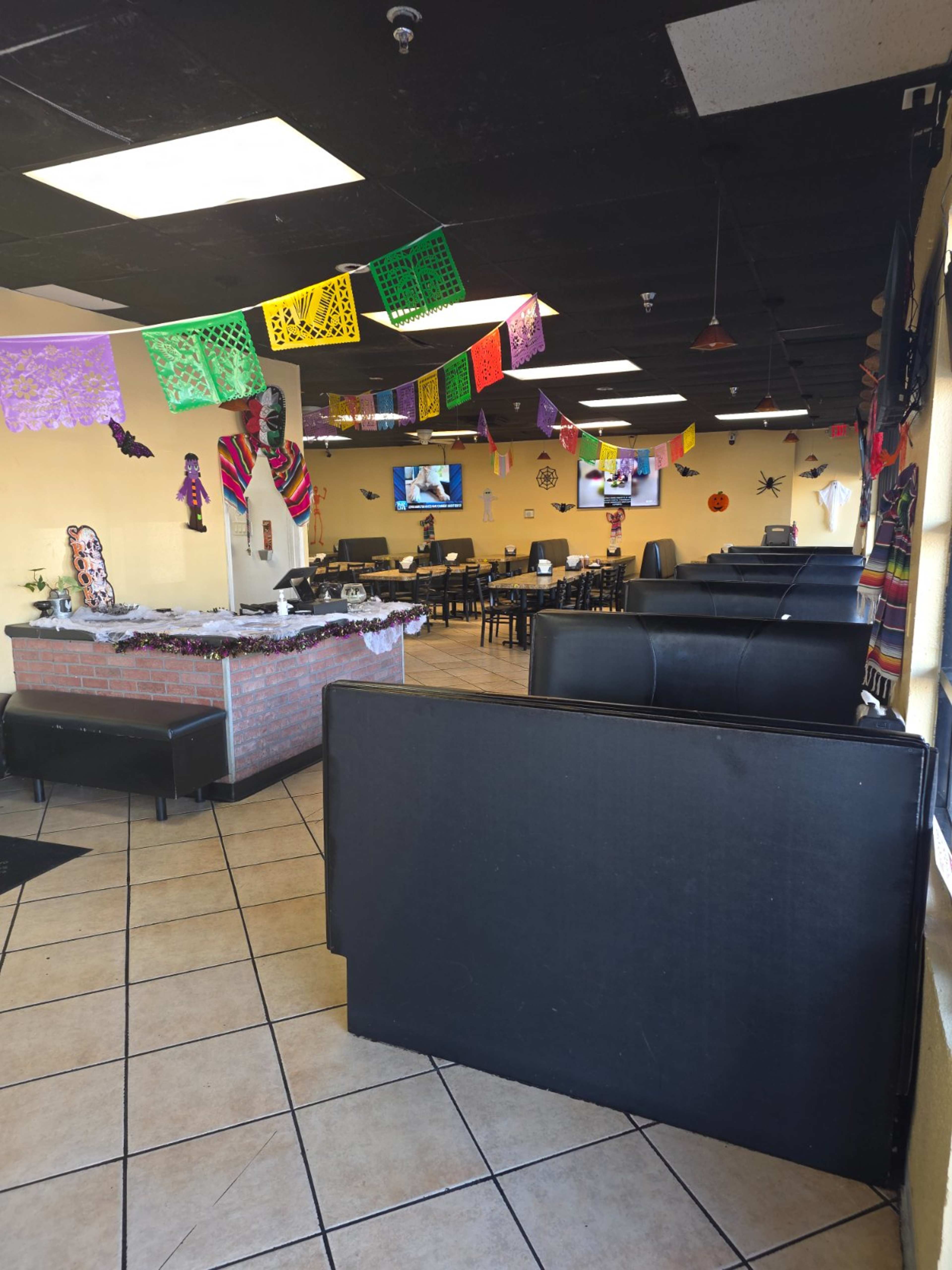 The image shows the interior of a restaurant featuring black seating, colorful papel picado decorations hanging from the ceiling, and tables arranged for dining.