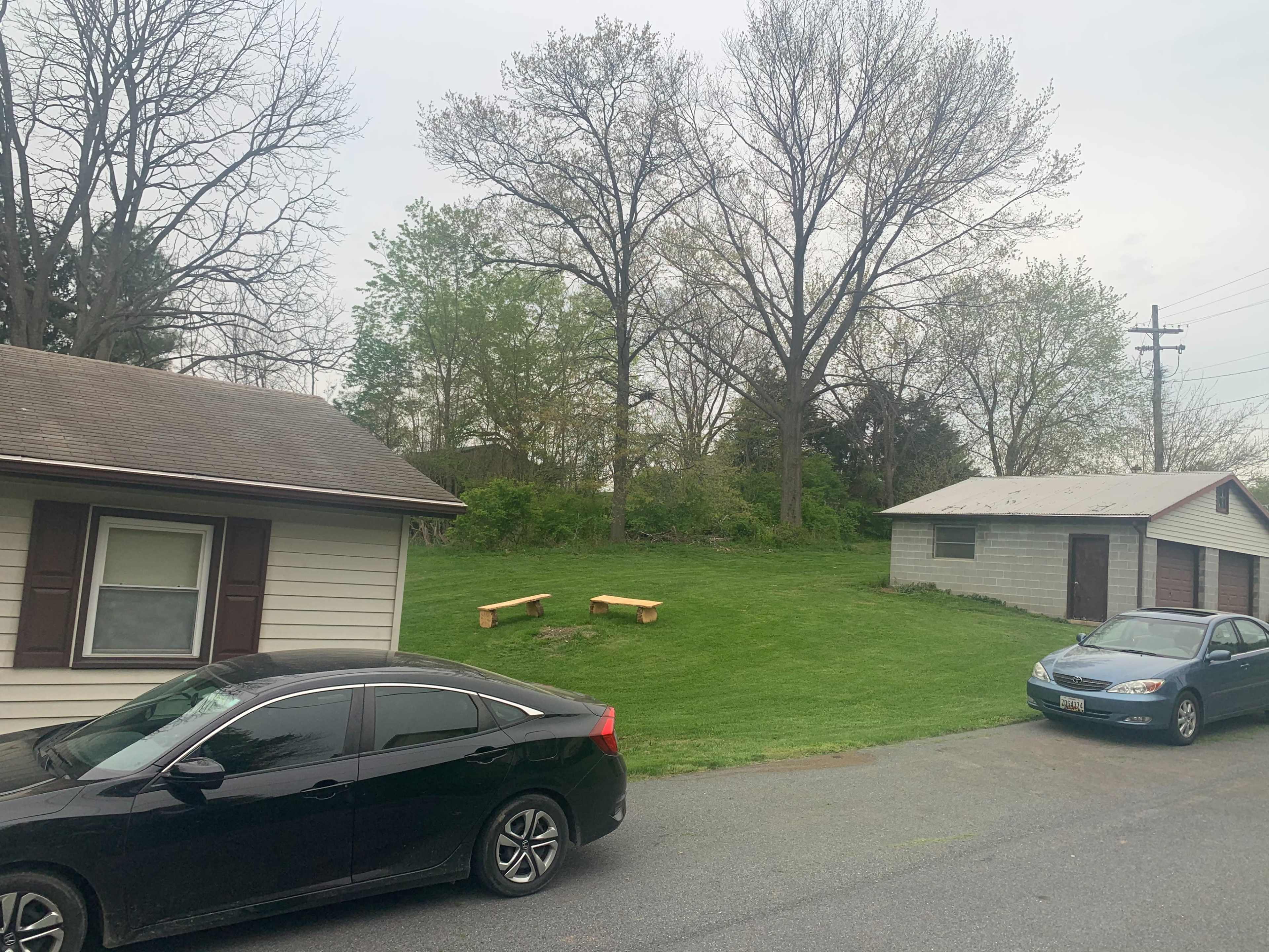 The image shows a grassy area with two wooden benches located between a house and a shed, with parked cars visible in the foreground.