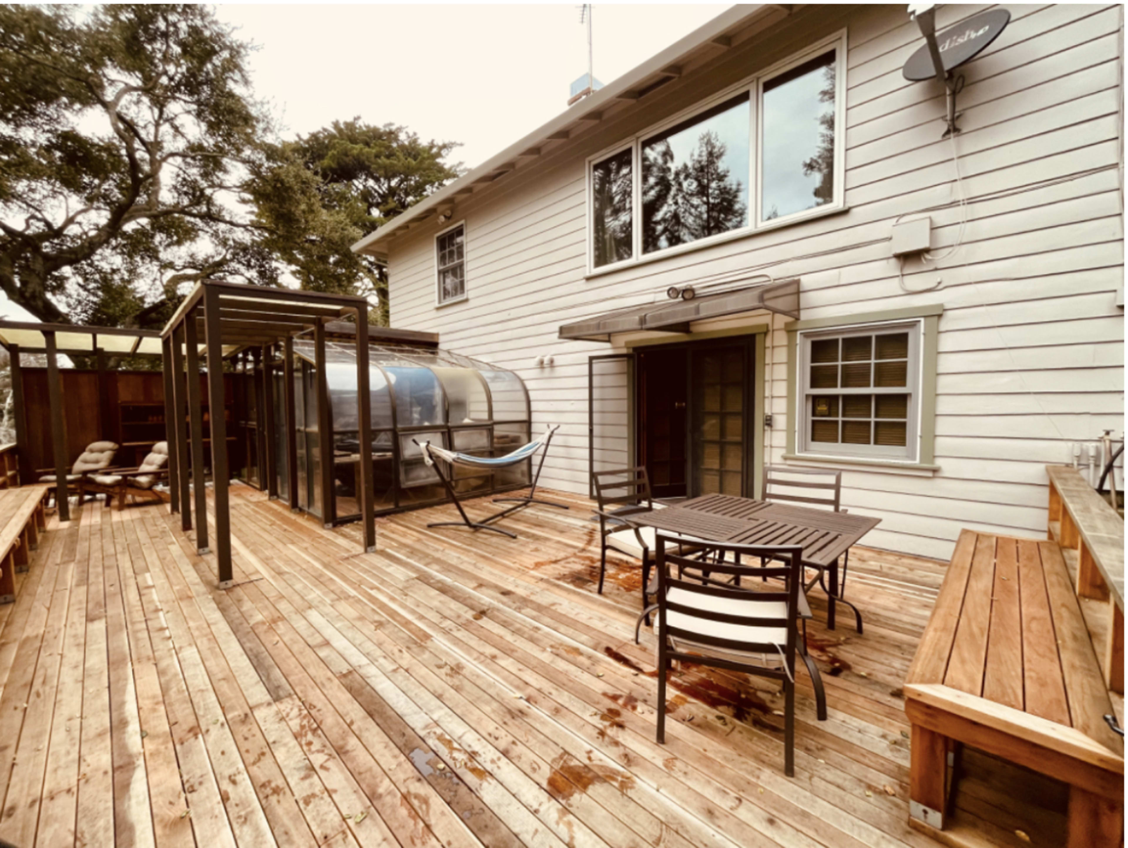 A wooden deck attached to a house, featuring a table with chairs, a hammock, and a clear cover structure.