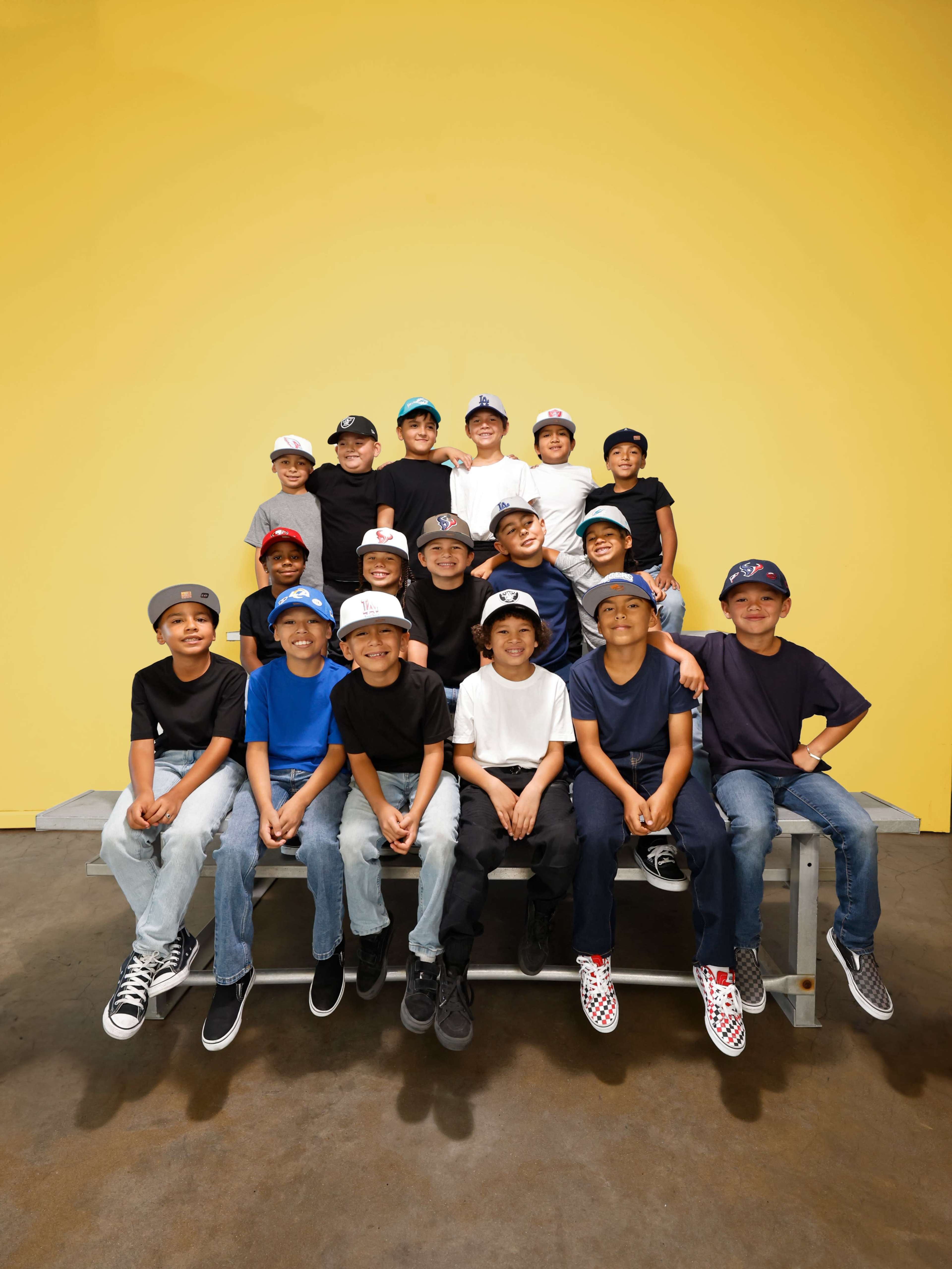 A group of fourteen children wearing various colored shirts and caps poses on a bench in front of a bright yellow wall.