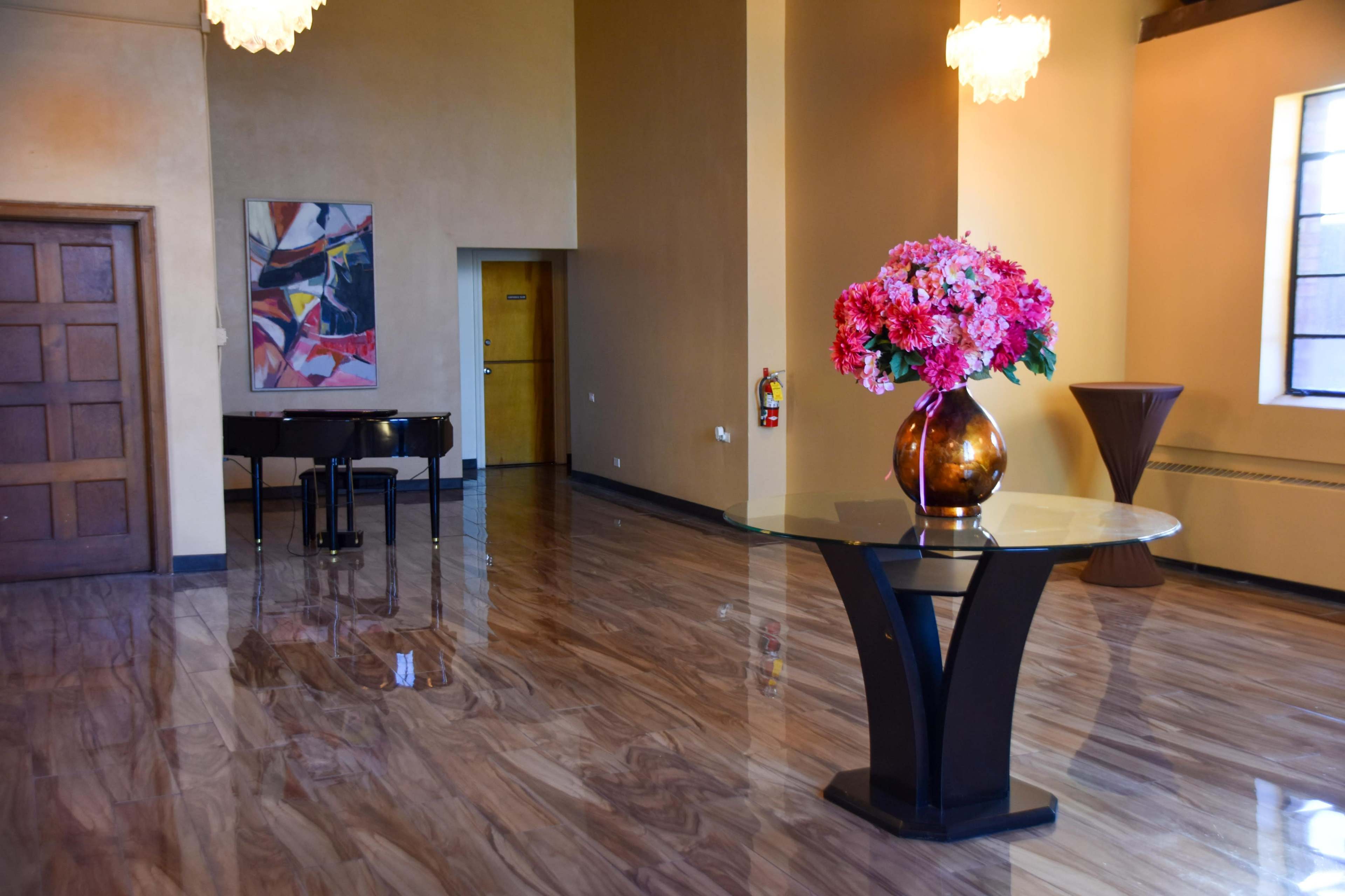 The image shows a spacious room with polished marble floors, featuring a grand piano, a round table with a floral arrangement, and a colorful abstract painting on the wall.