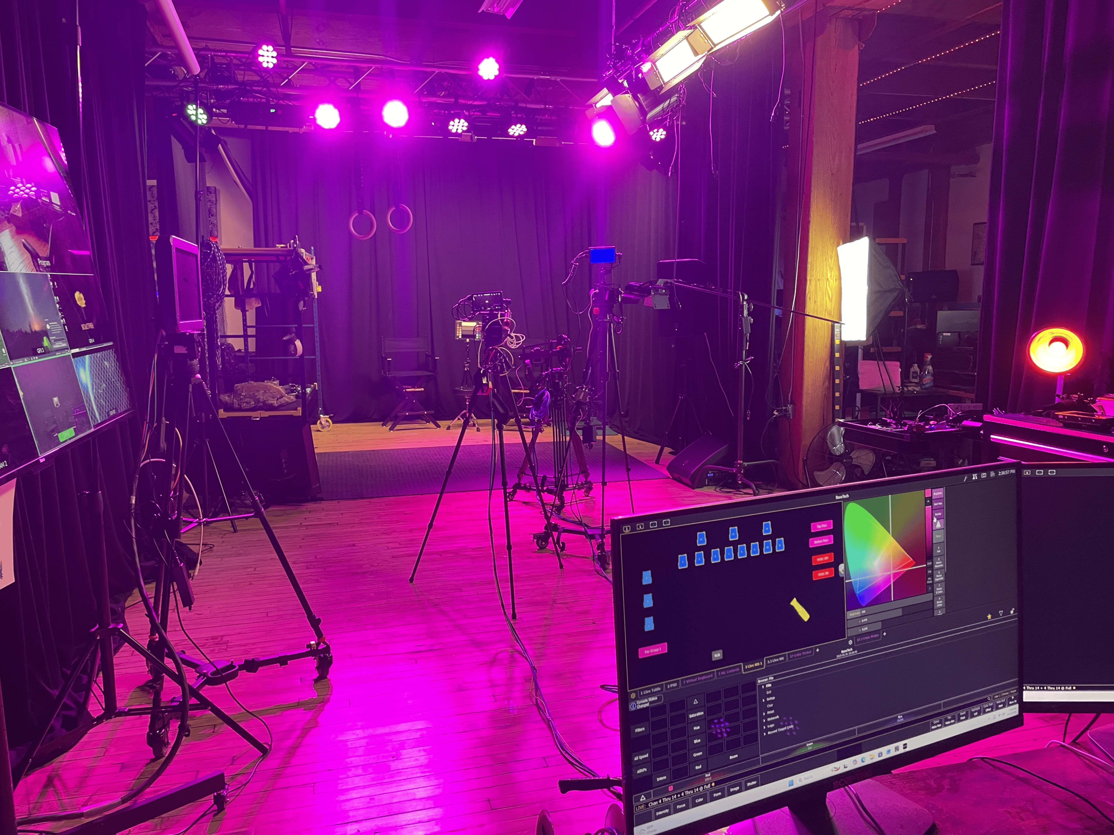 The image shows a dimly lit studio setup with multiple cameras, lighting equipment, and computer monitors arranged on the floor.