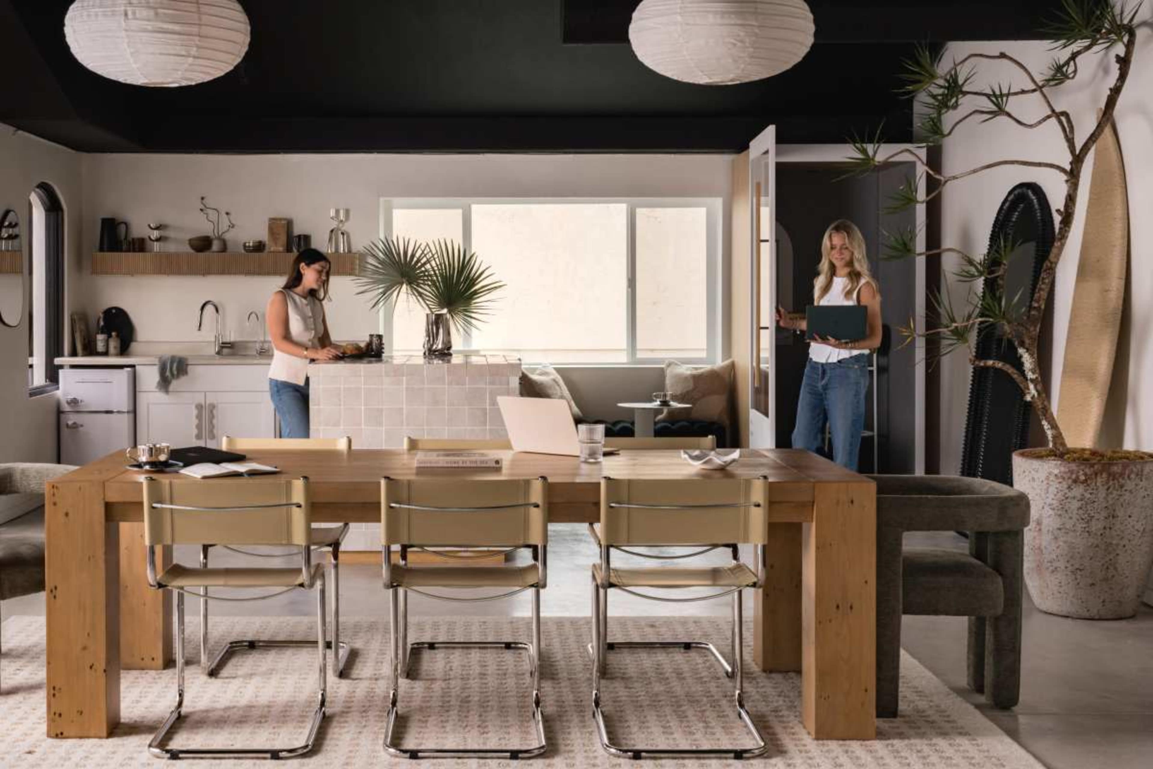 A modern office space features a wooden dining table surrounded by four metal chairs, where two women engage in work-related activities, with large windows and plants enhancing the aesthetic.