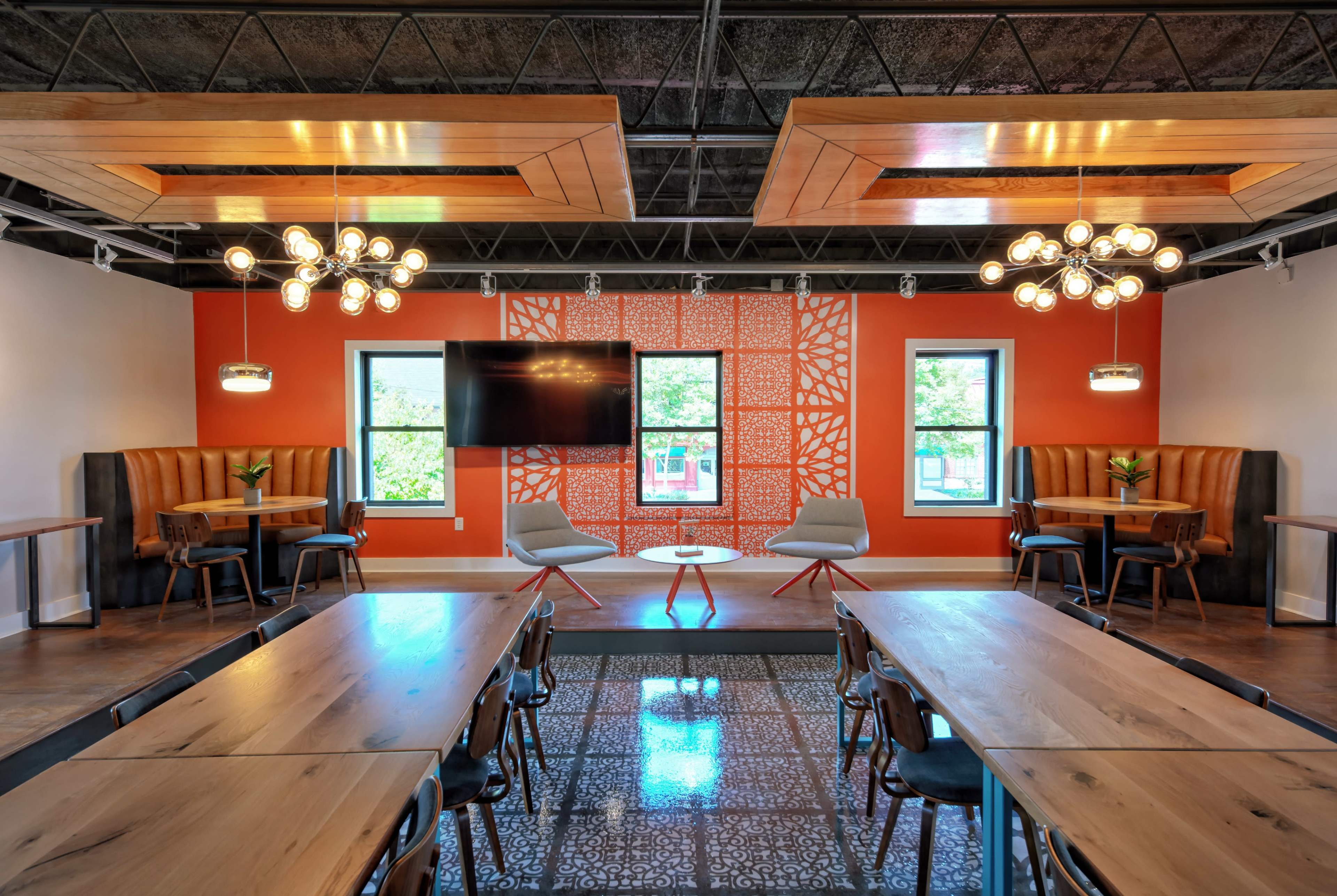 The image shows a modern meeting room with wooden tables, stylish seating areas, and an orange accent wall featuring geometric patterns.
