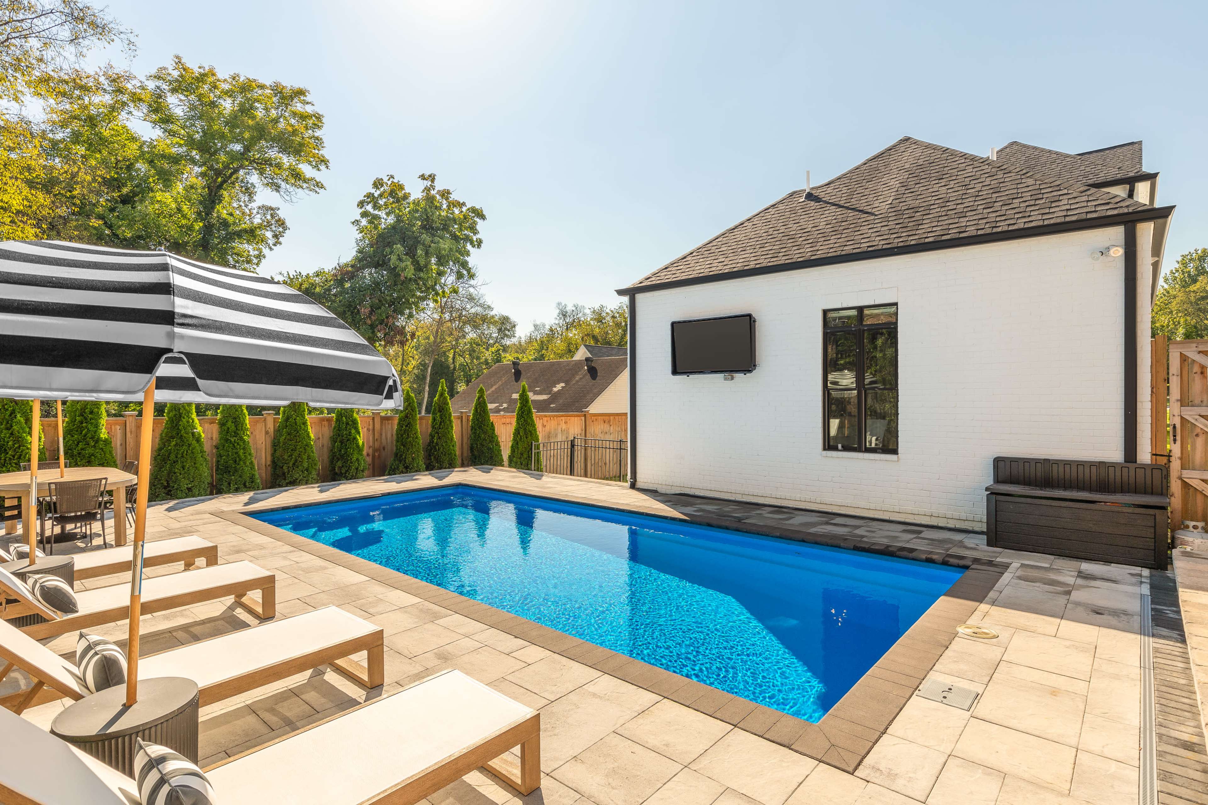 The image shows a residential backyard featuring a rectangular swimming pool surrounded by lounge chairs and an umbrella, with a corner of the house visible in the background.