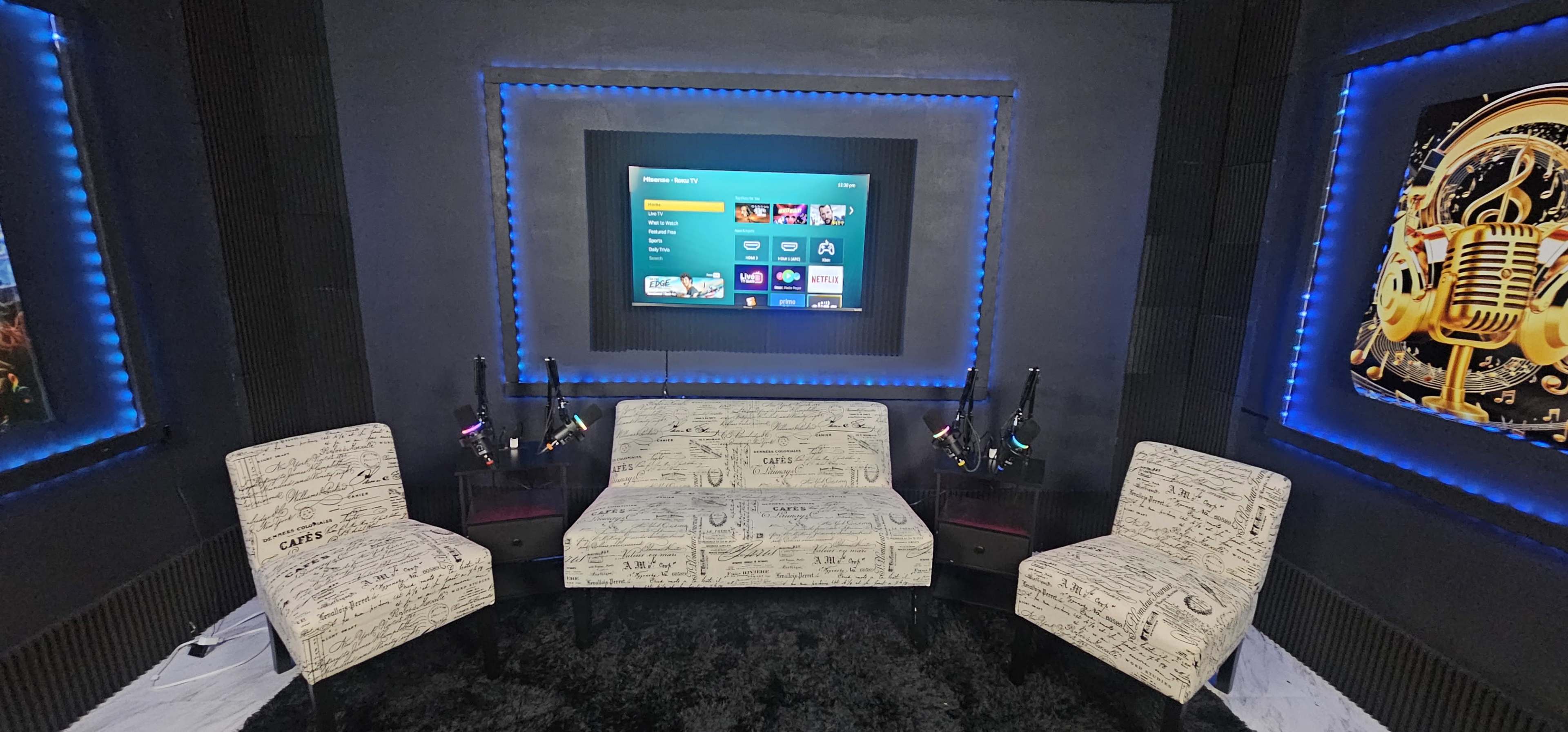 The image shows a modern lounge area with a black carpet, two patterned chairs, and a central couch facing a wall-mounted screen, all accented by blue LED lighting.