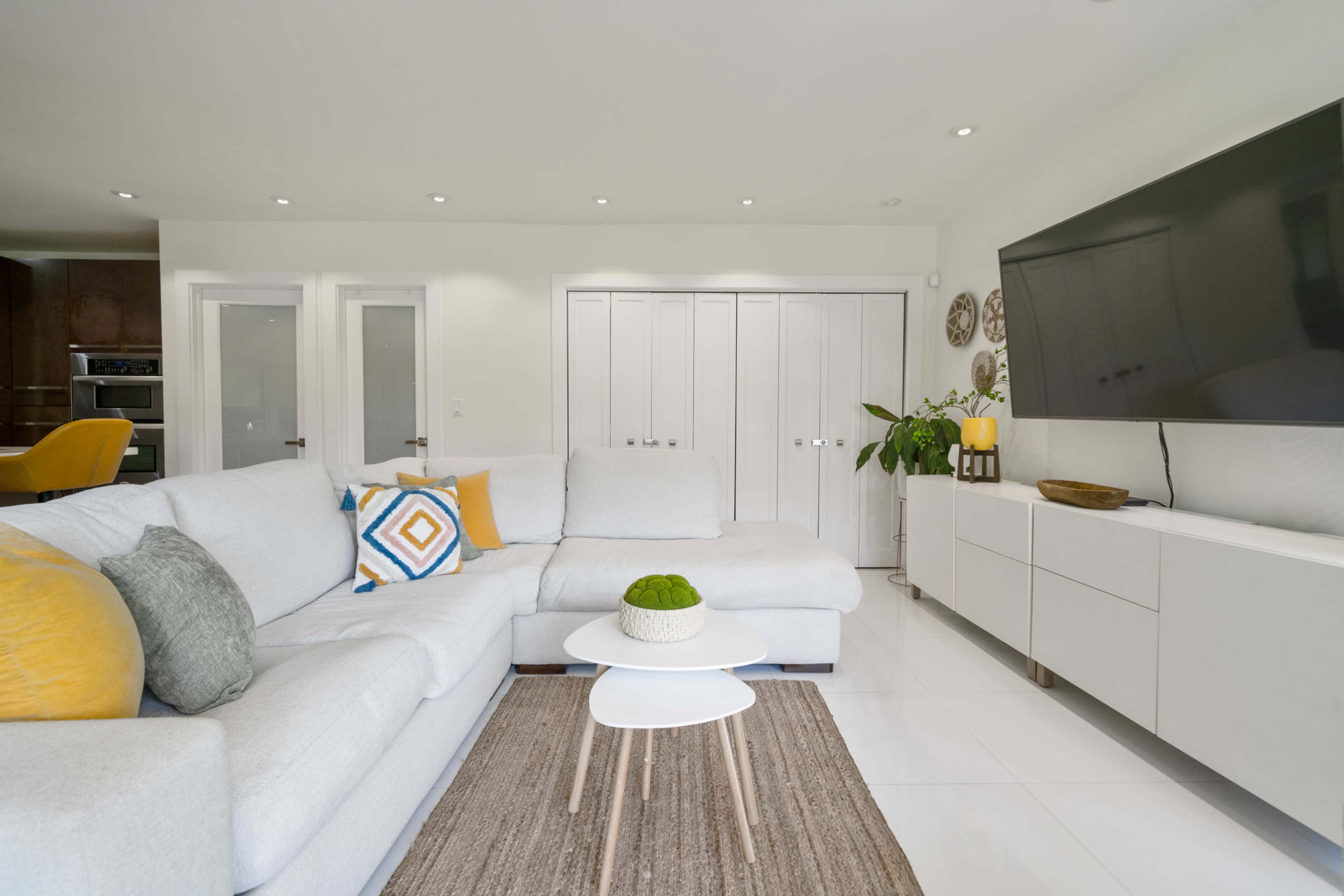 A light-filled living room features a large gray sectional sofa, a round coffee table, and a wall-mounted television, with minimalist decor and plants in the background.