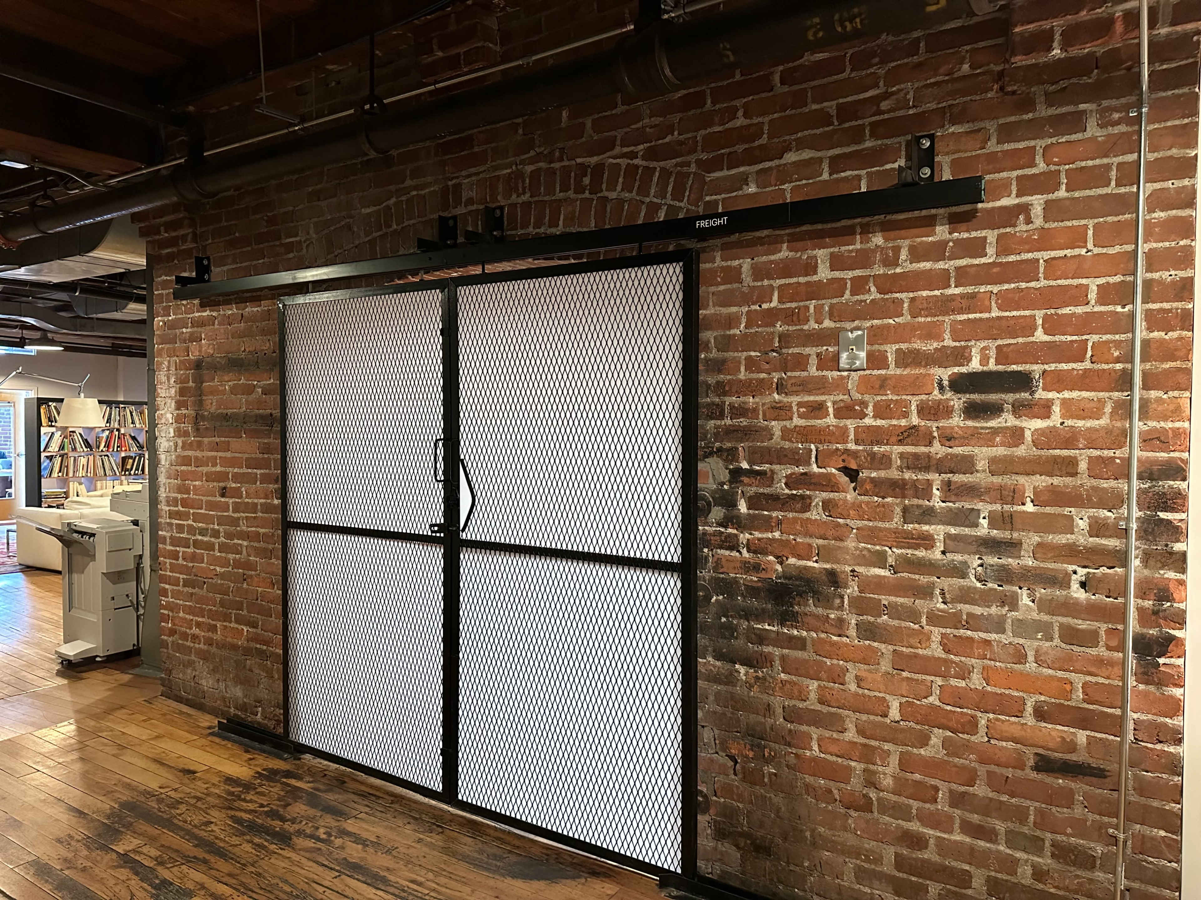A black metal sliding door with a mesh design installed in front of a brick wall in an interior space.