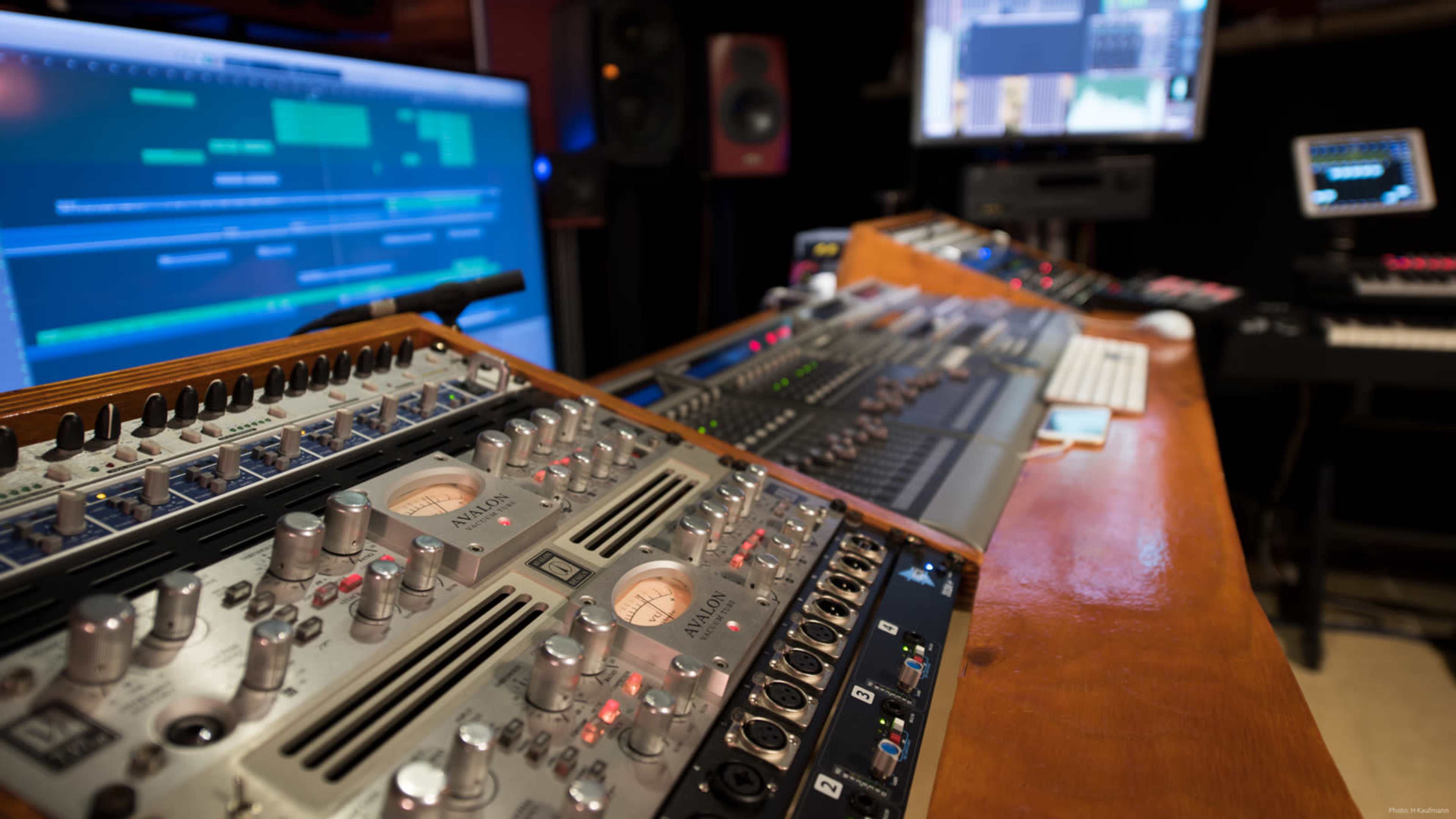 A close-up view of a music production studio featuring various audio mixing equipment and multiple computer screens displaying software.