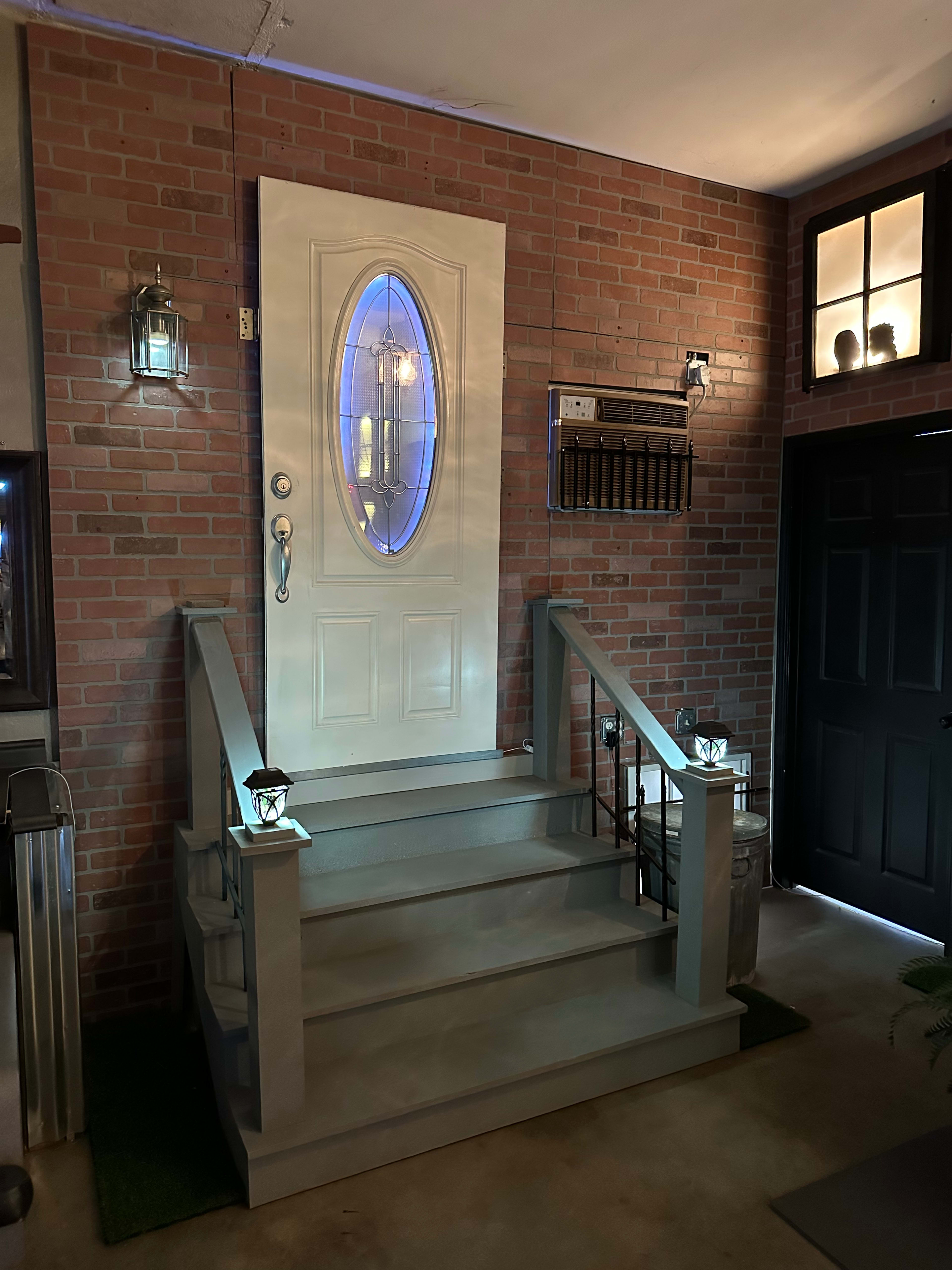 A white door with an oval window is set in a brick wall, surrounded by stairs and decorative lanterns.