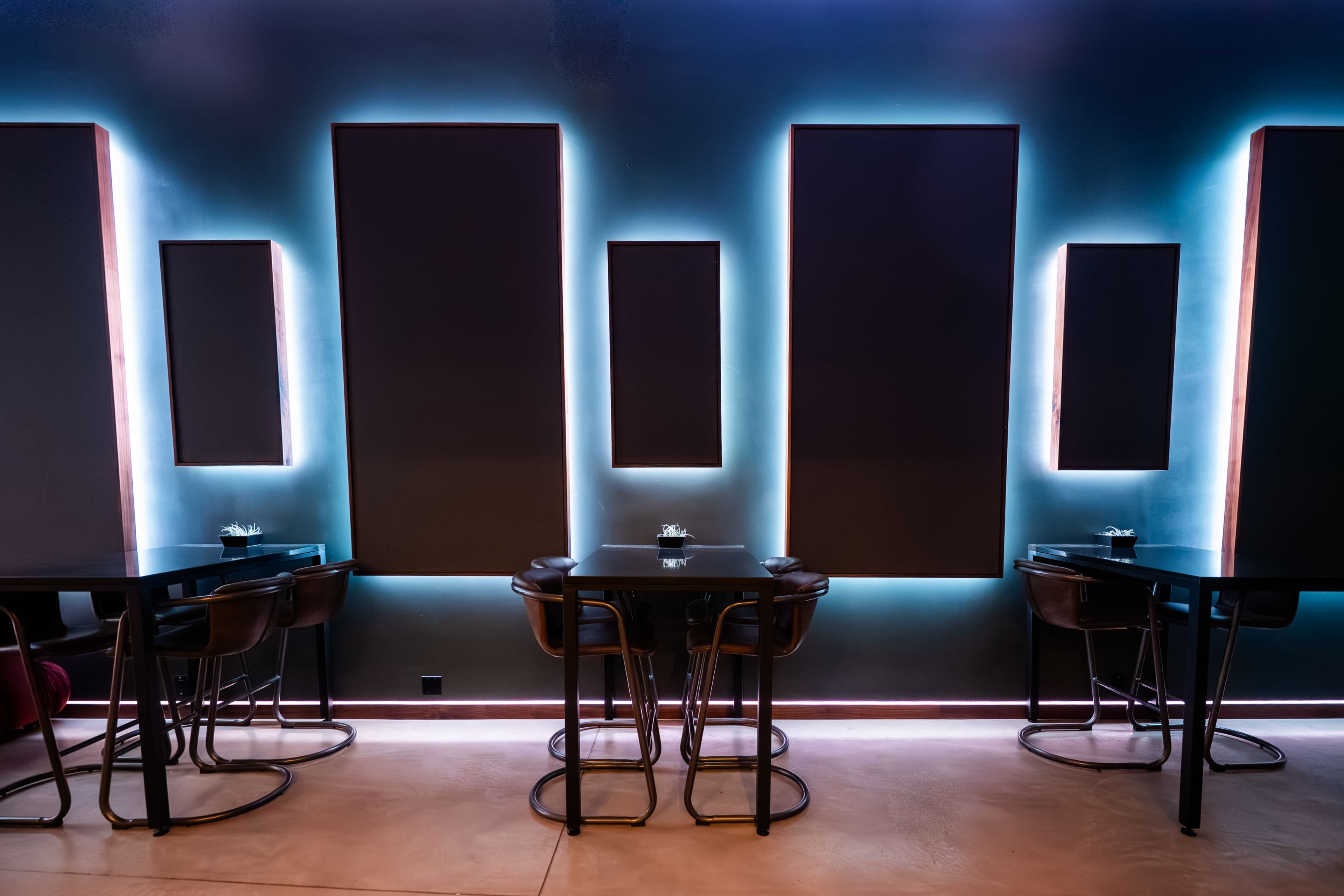 The image shows a modern restaurant interior with illuminated wall panels and black tables accompanied by high-backed chairs.