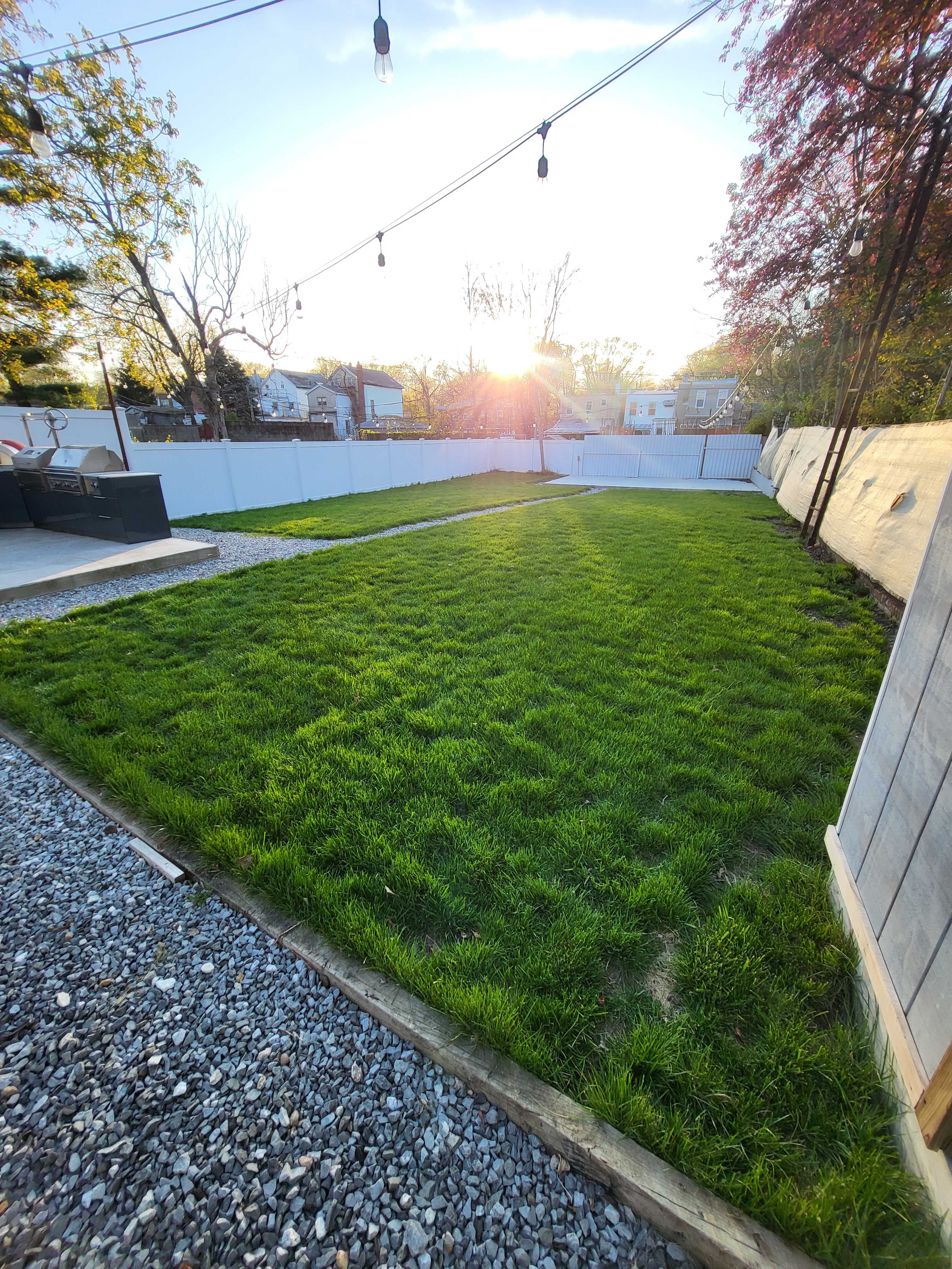 A backyard with freshly grown grass, a gravel pathway on one side, and the sun setting in the background.
