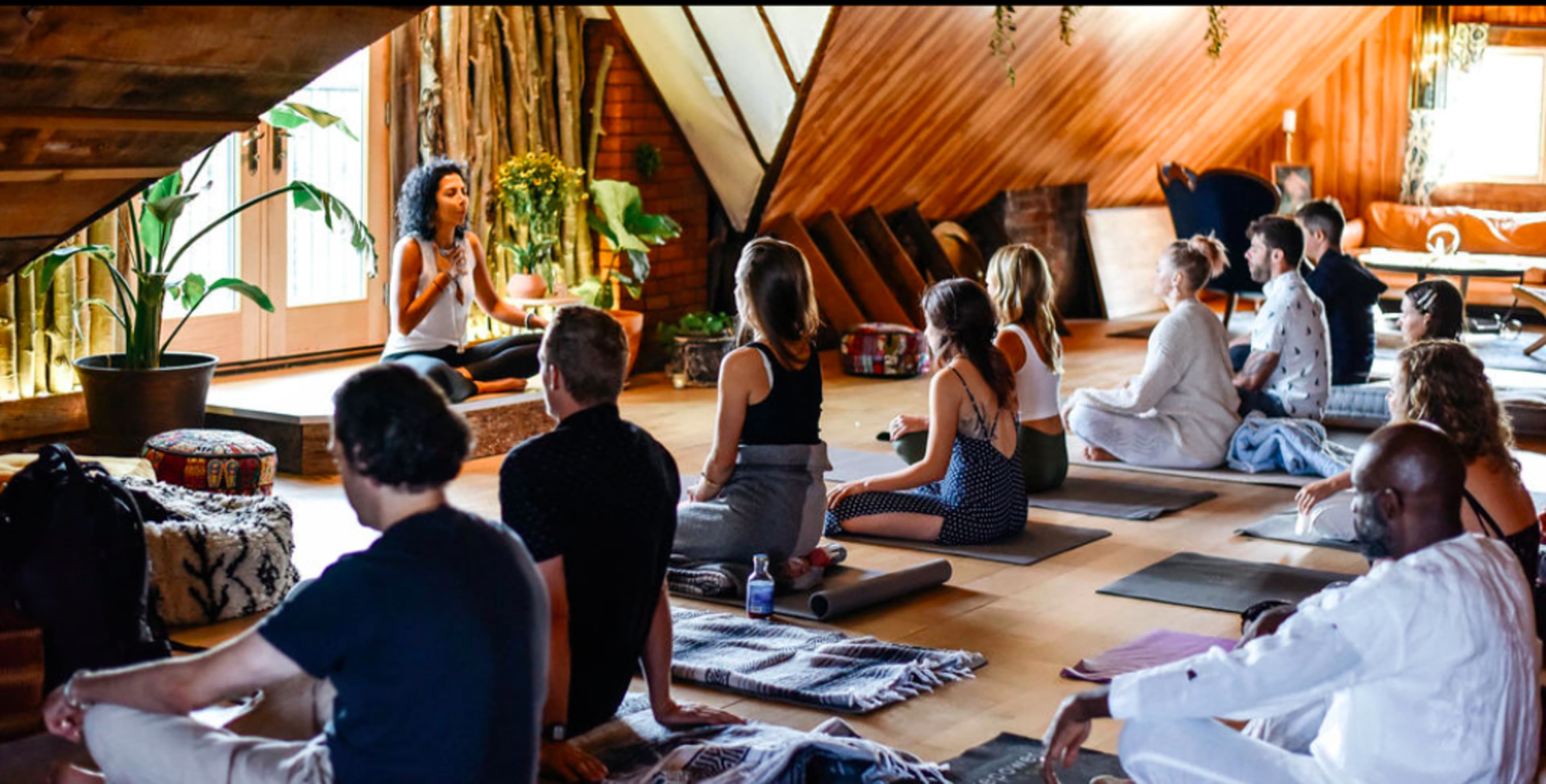 A group of people are seated on yoga mats in a warmly lit studio, listening to an instructor leading a session.
