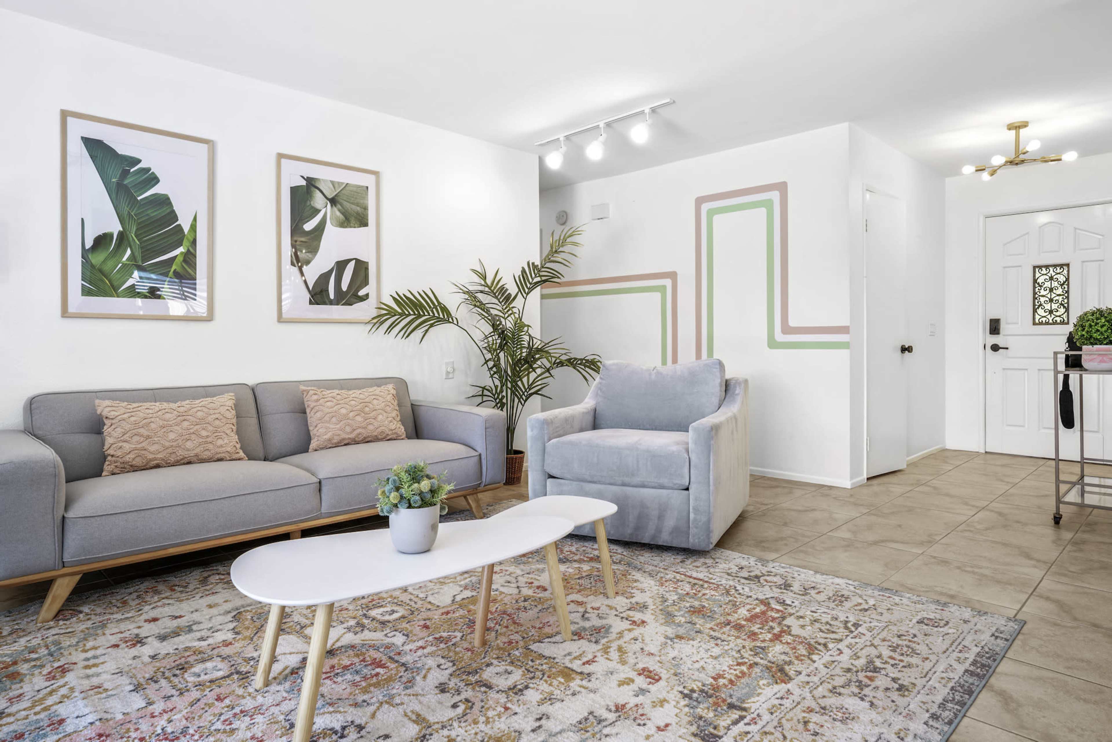 A living room features a light gray sofa, a matching armchair, a coffee table, potted plants, and framed botanical prints on the walls.