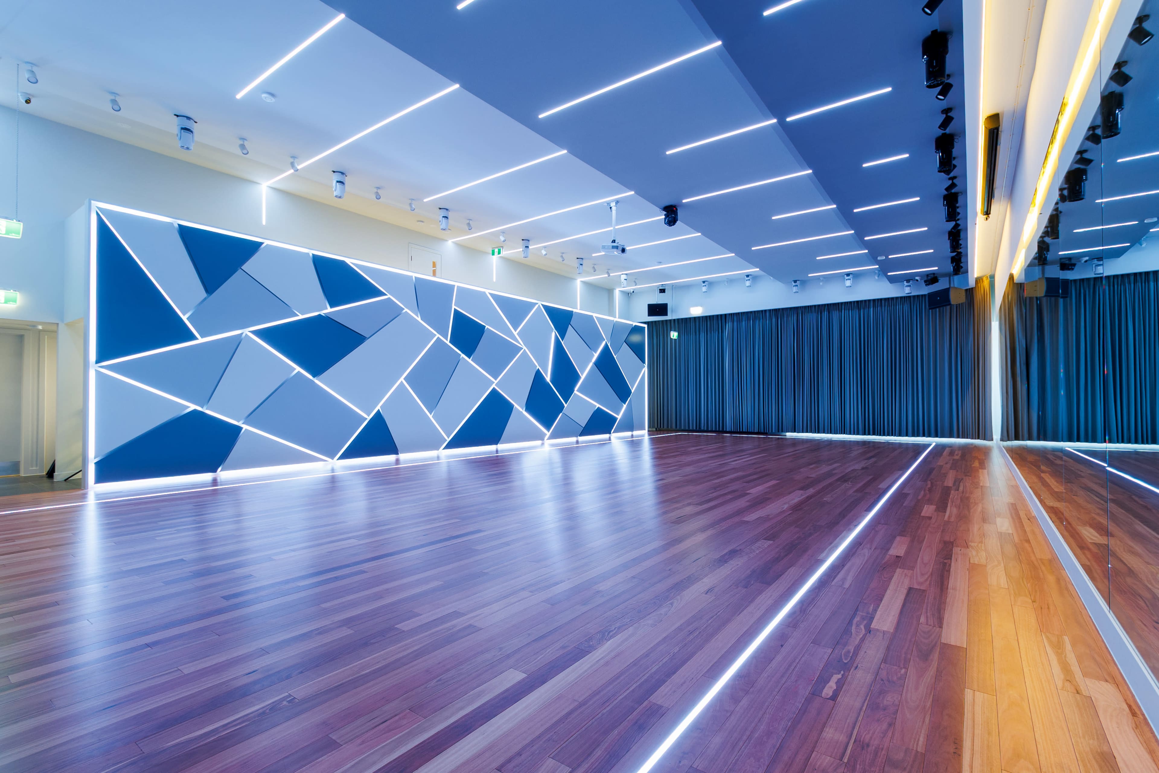 The image shows a modern dance studio with a geometric blue and white accent wall, hardwood floors, and bright overhead lighting.