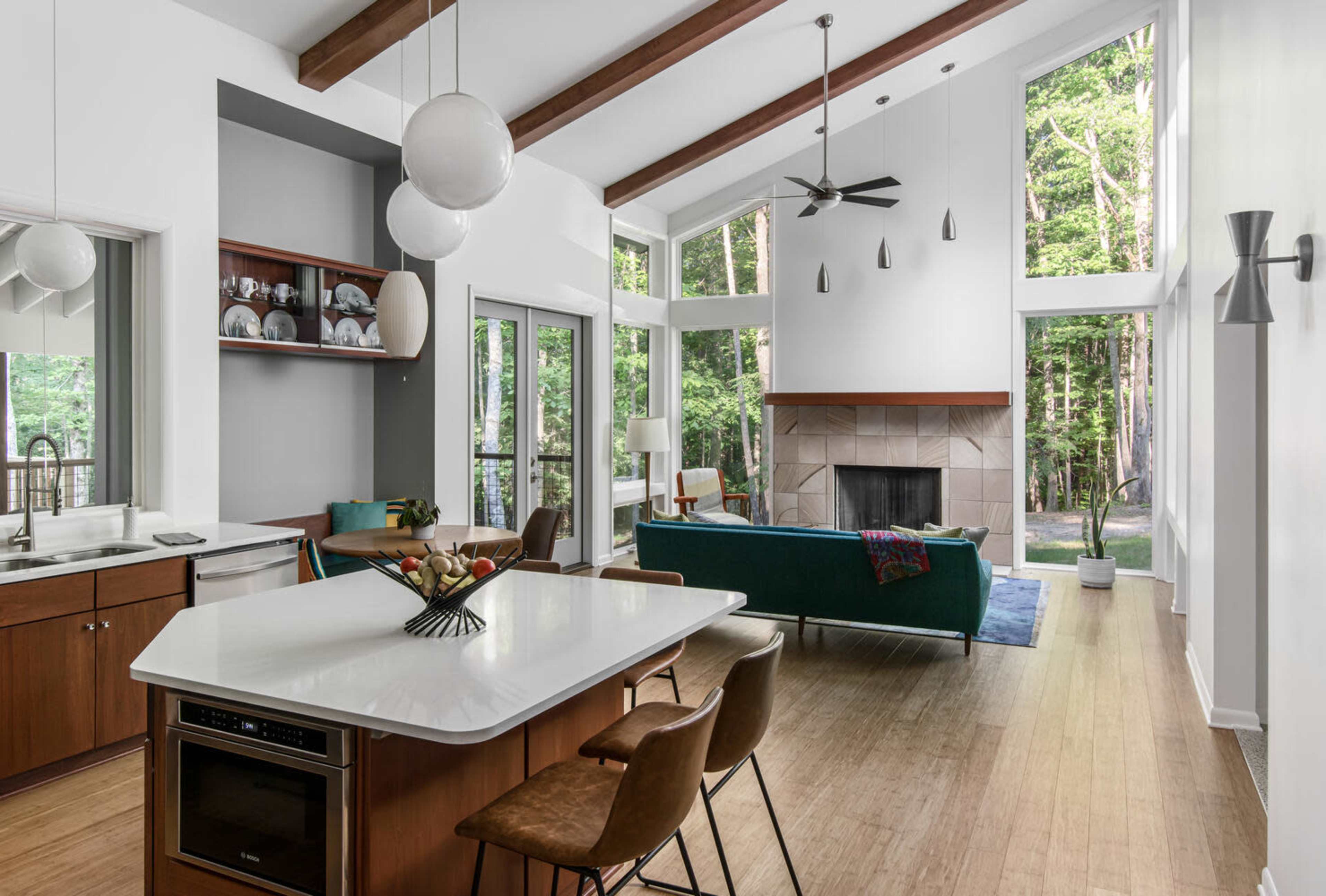 The image shows a modern kitchen and living area with high ceilings, large windows, wooden beams, and a fireplace.