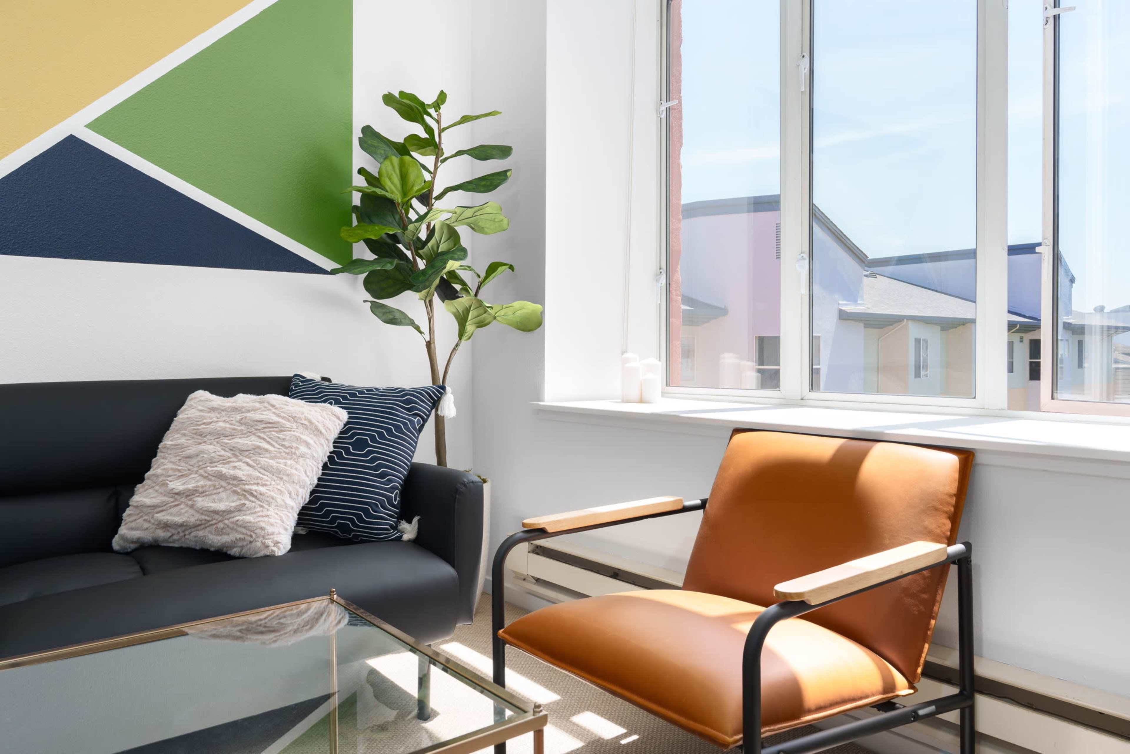 A modern living space with a black sofa, a brown armchair, a potted plant, and large windows allowing natural light to enter.