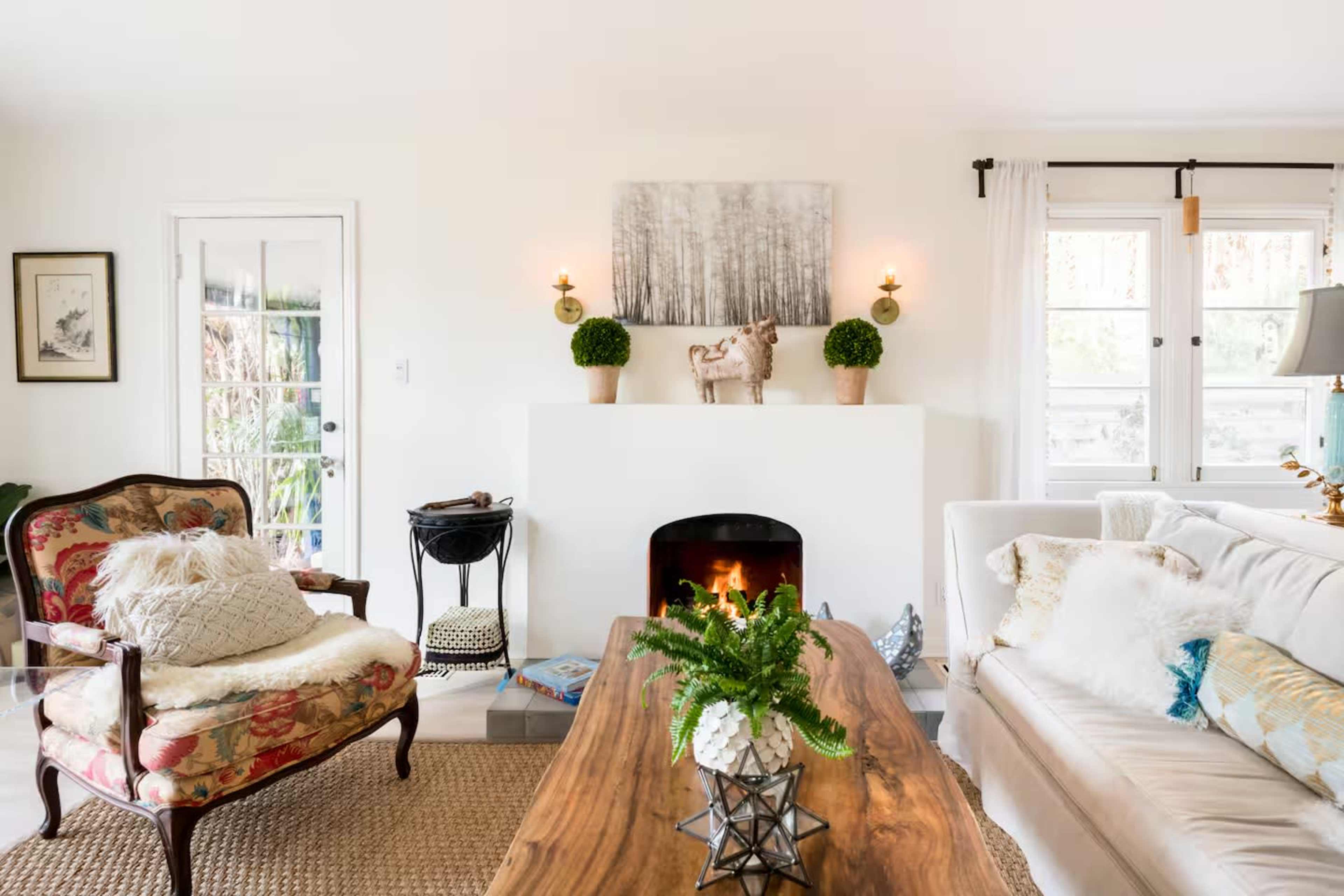 The living room features a white color scheme with a cozy sofa, a floral armchair, a wooden coffee table, and decorative wall art, centered around a fireplace.