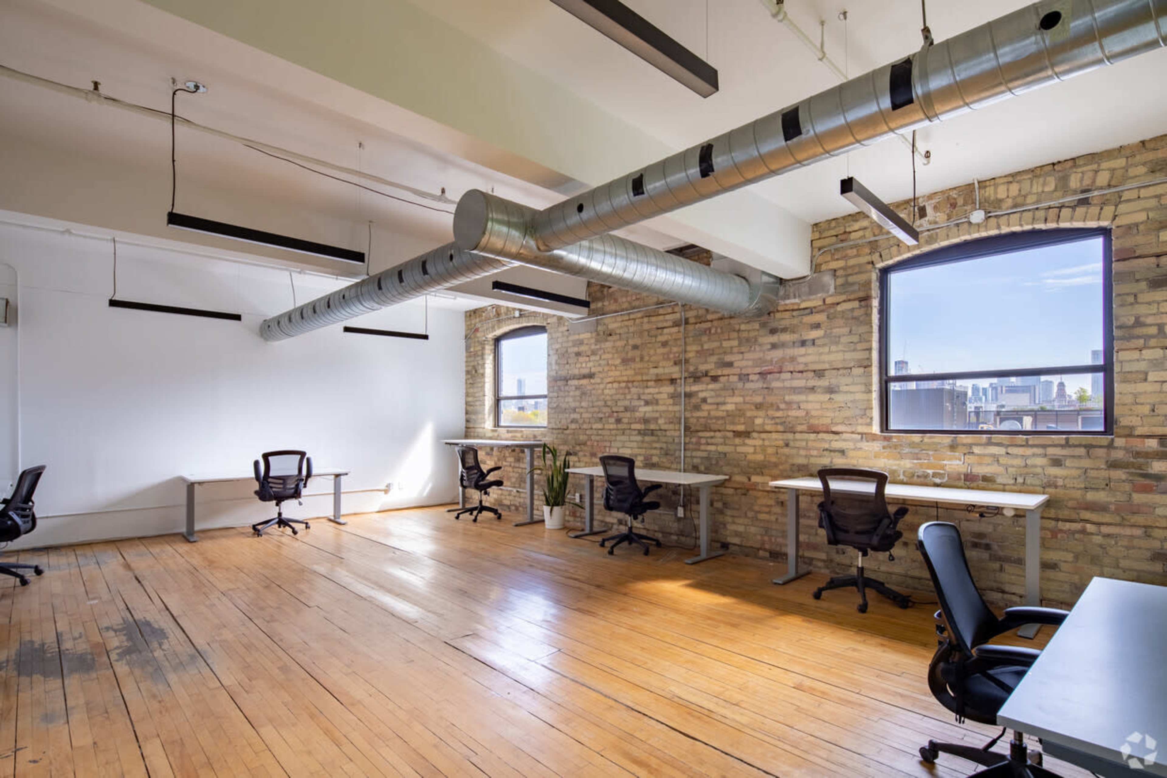 The image shows an empty office space with wooden floors, large windows, and exposed ductwork along the ceiling.