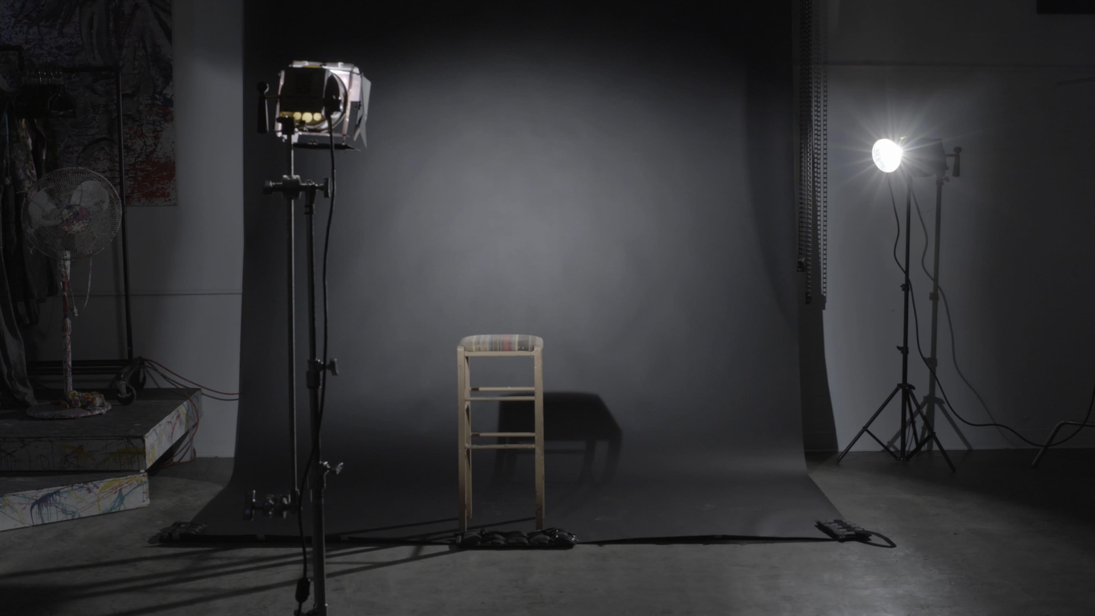 A wooden chair is positioned in front of a dark backdrop, illuminated by two lights on either side.