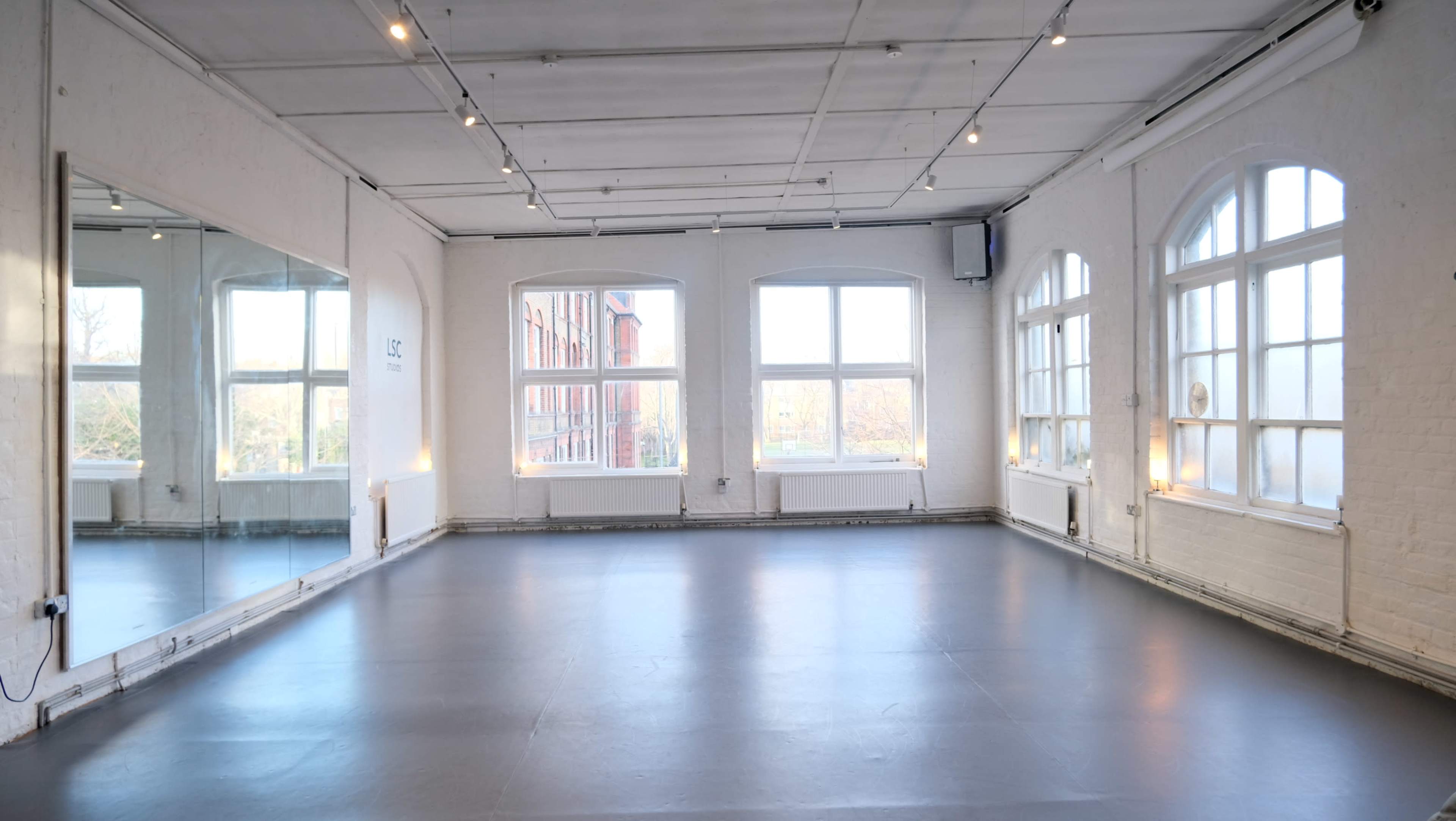 The image shows a bright, spacious room with large windows, a mirrored wall, and a smooth floor, designed for activities such as dance or yoga.