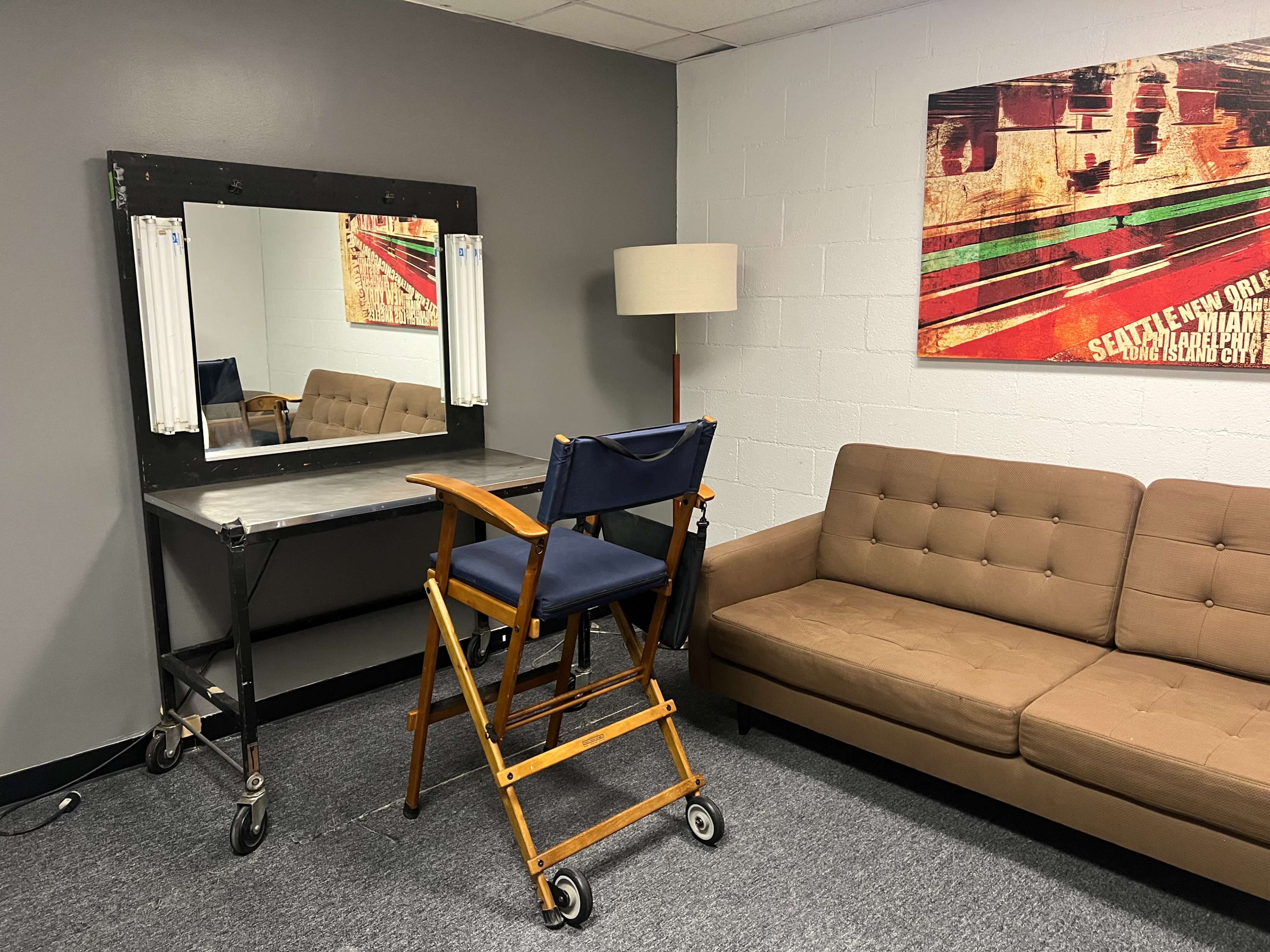 A small room featuring a director's chair in front of a lit makeup mirror, a lamp, and a couch against the wall with a colorful artwork above it.