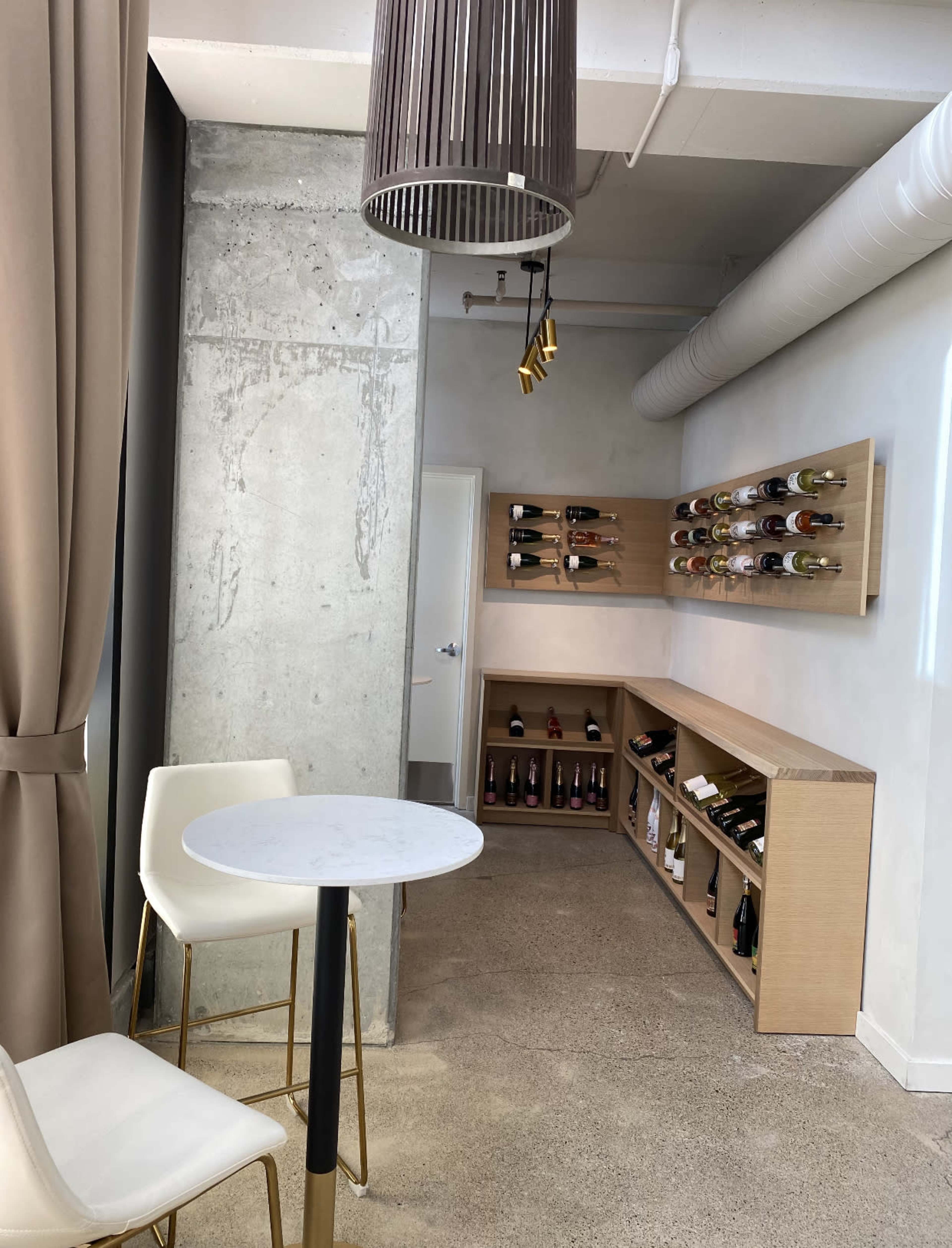 A modern interior with a small round table and two white chairs, concrete walls, and a wine display along the wall.