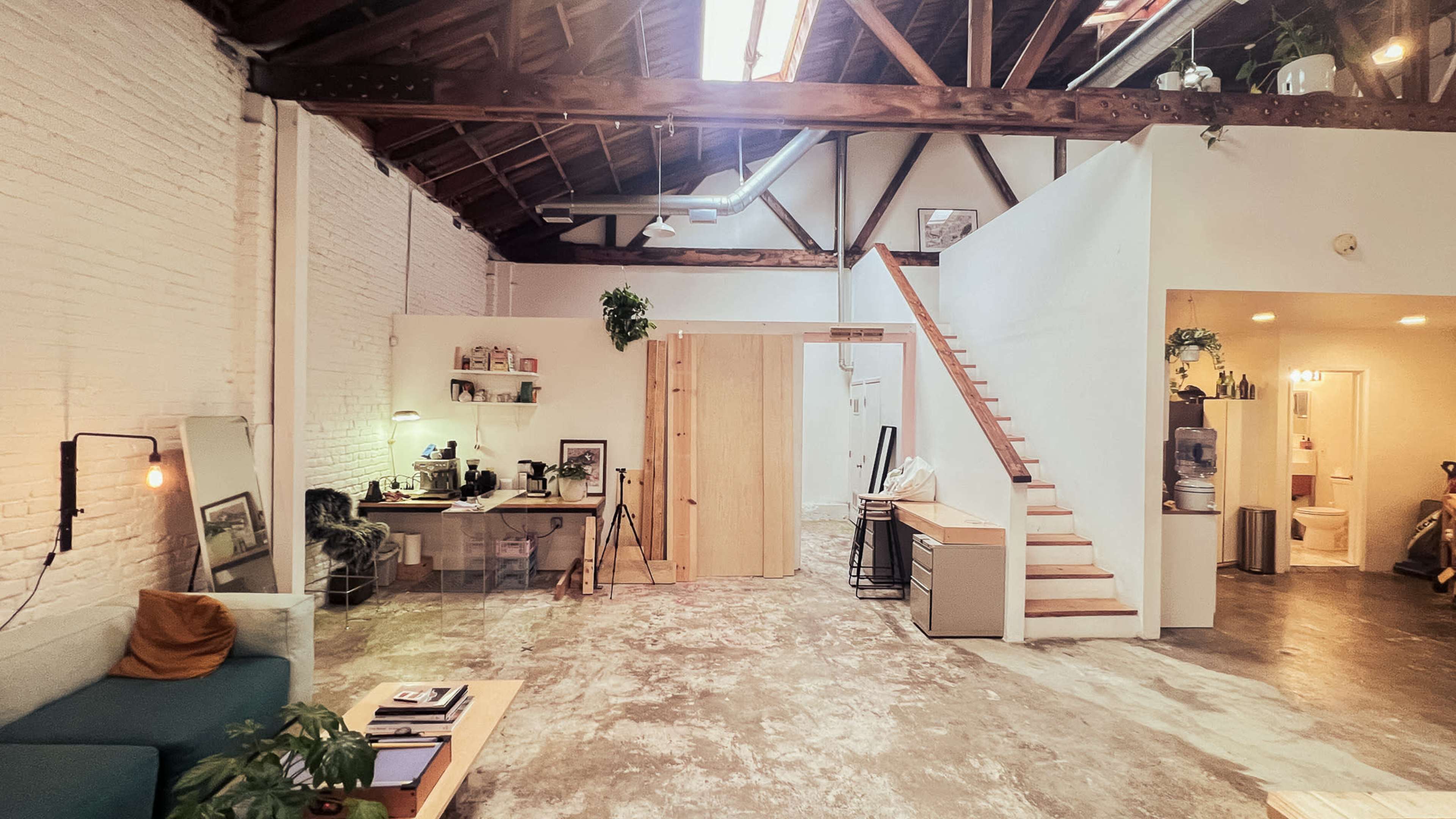 The image shows an open-concept loft with a wooden staircase, a workspace with a desk and chair, and a small bathroom area in the background.