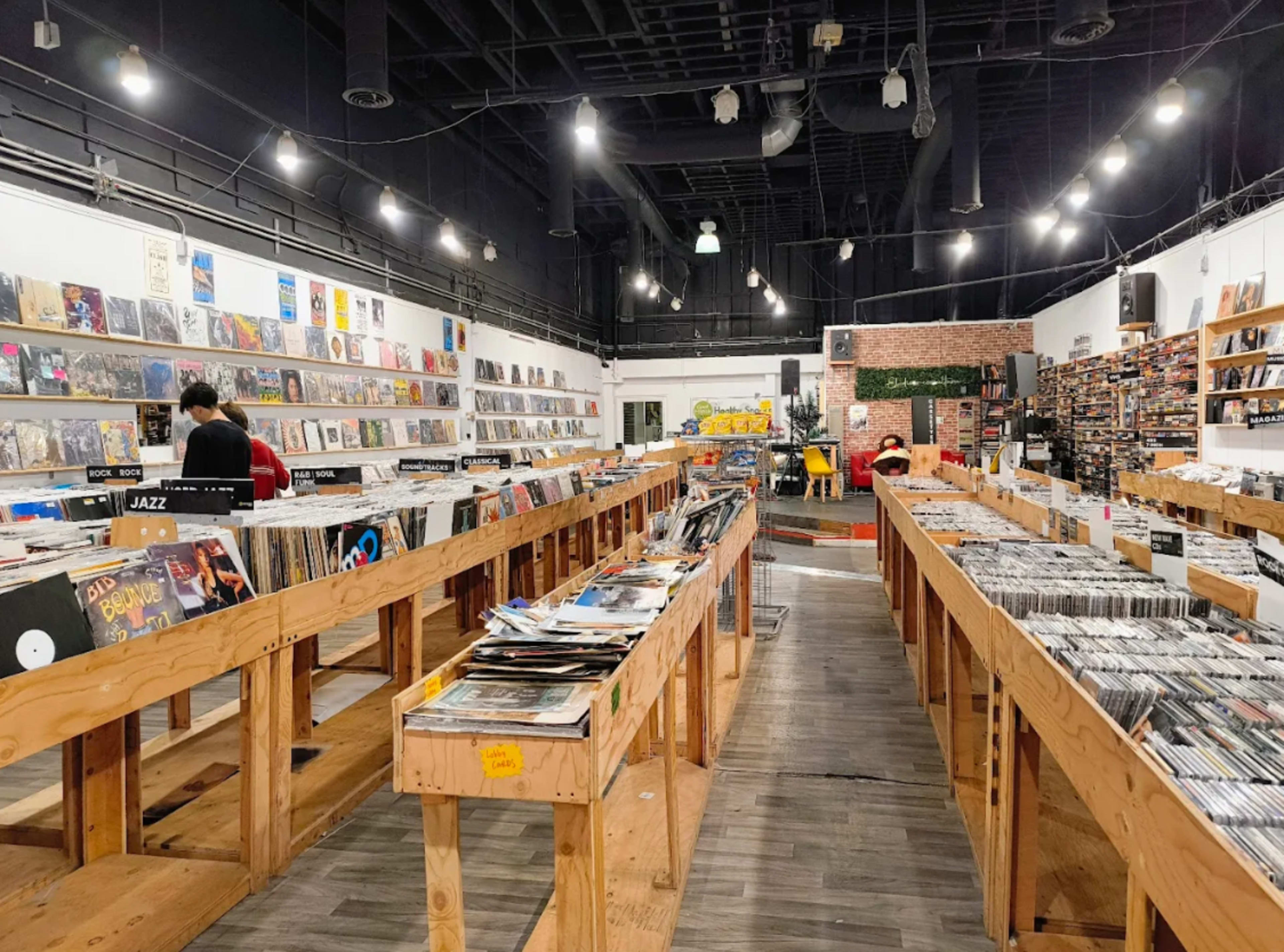 The image shows an interior of a record store with wooden tables lined with vinyl records and shelves filled with albums along the walls.