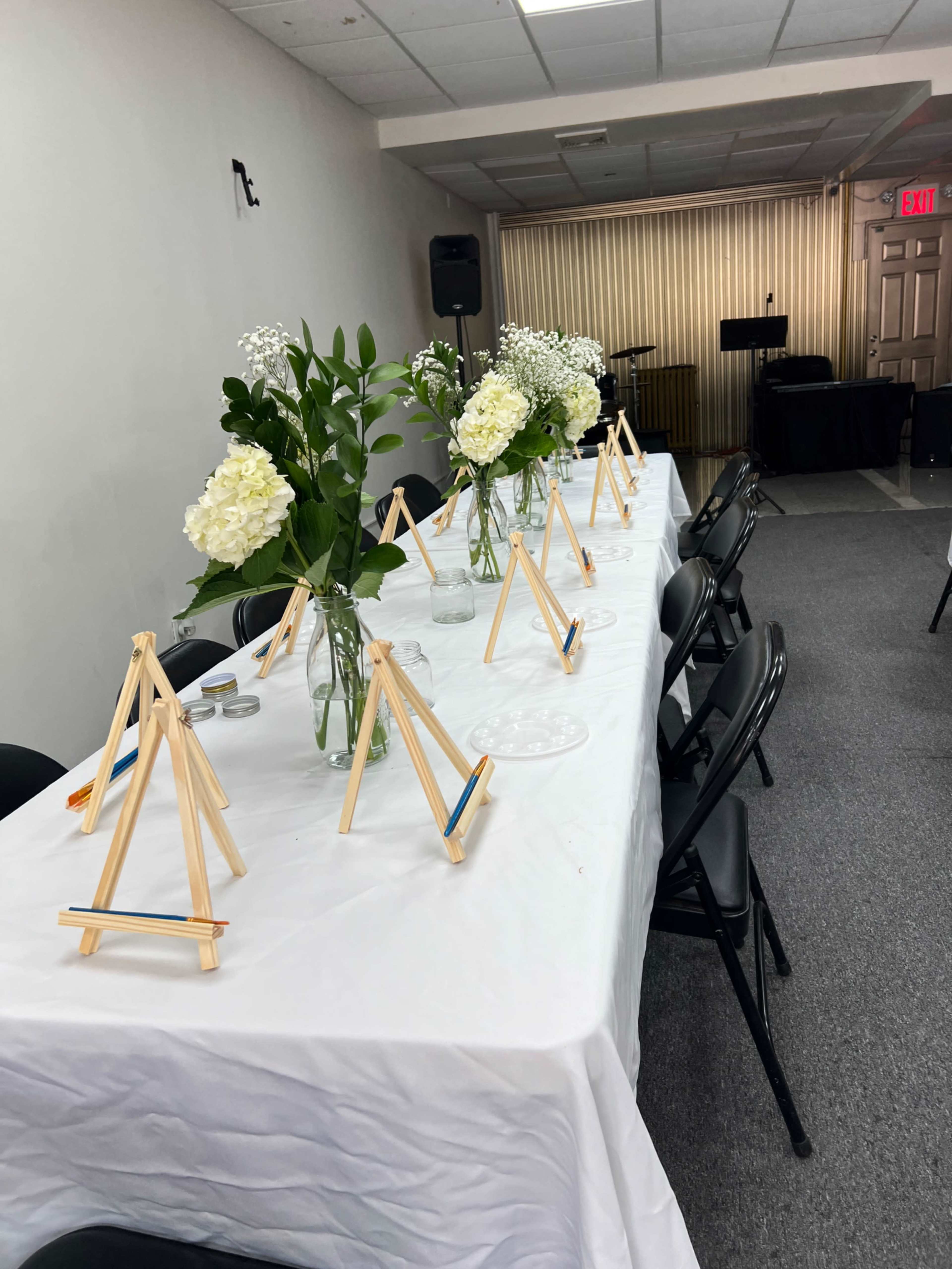 A long table covered with a white cloth is set up with small easels and vases of flowers in a room that appears to be prepared for an event.