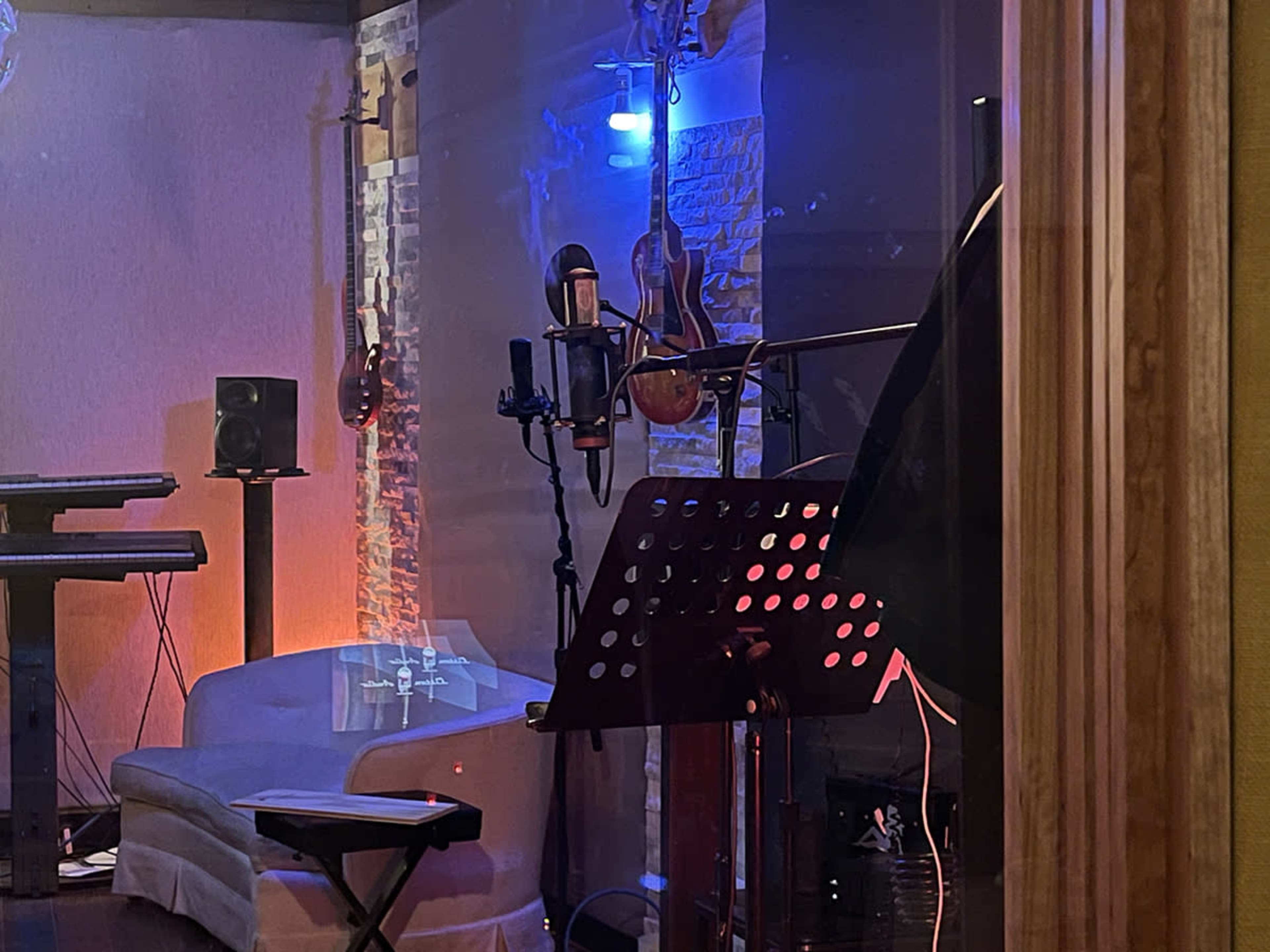 The image shows a recording studio with a microphone setup, musical instruments hanging on the wall, and a small white couch.