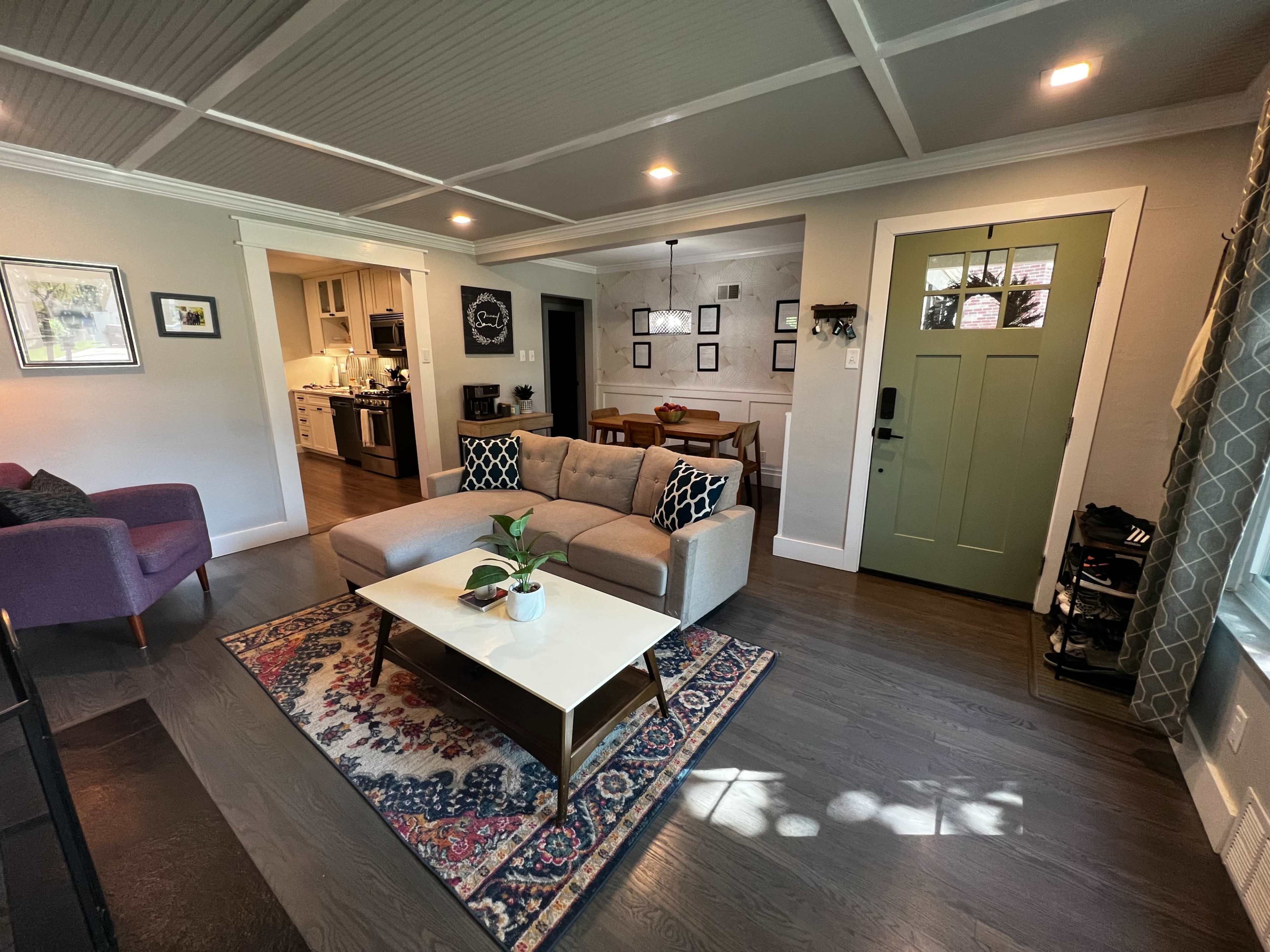 A cozy living room features a gray sectional sofa, a coffee table with a small plant, and a green front door, with a dining area visible in the background.