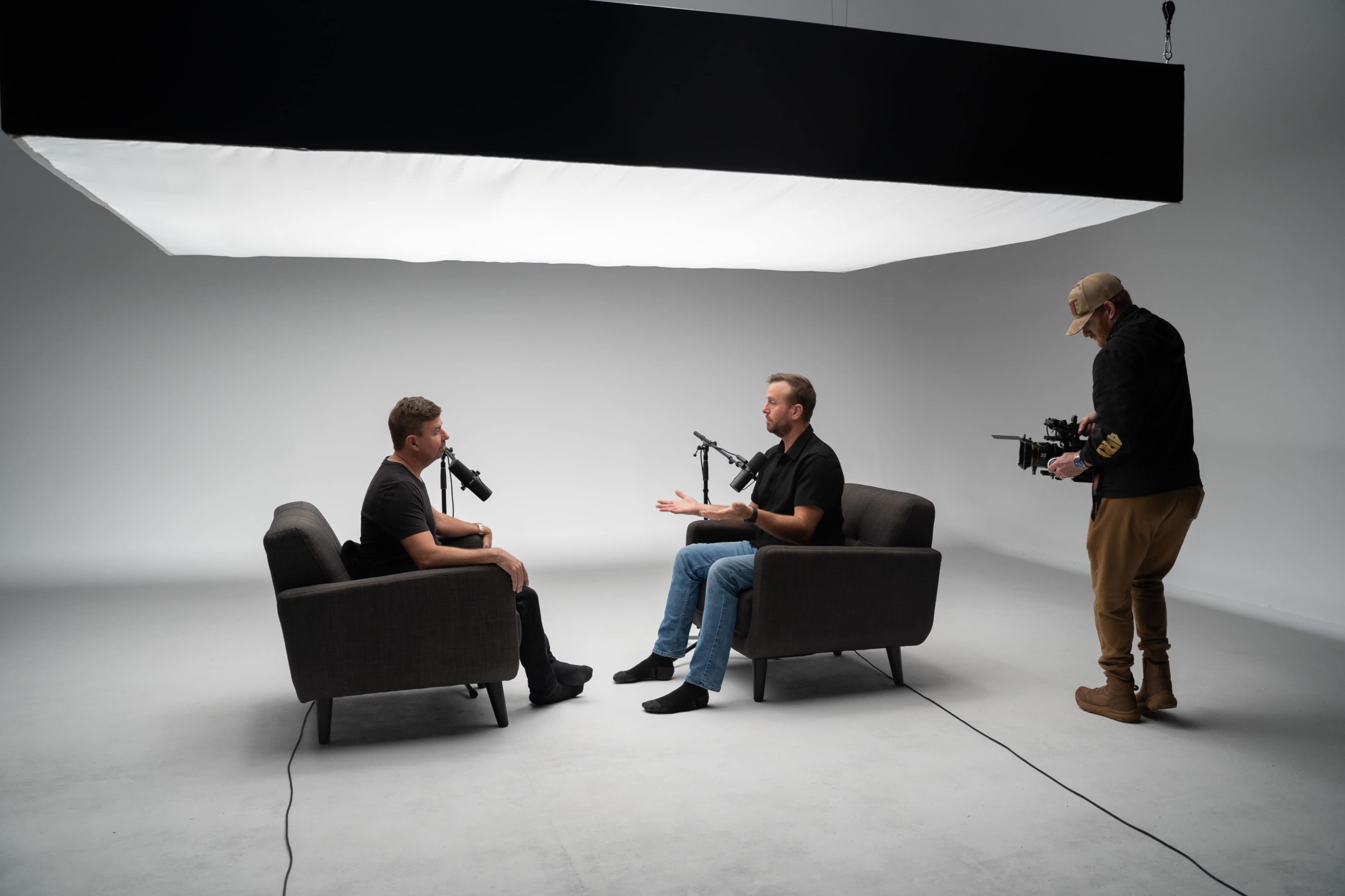Two men sit in chairs facing each other in a minimalist studio, while a third man records the conversation with a camera.