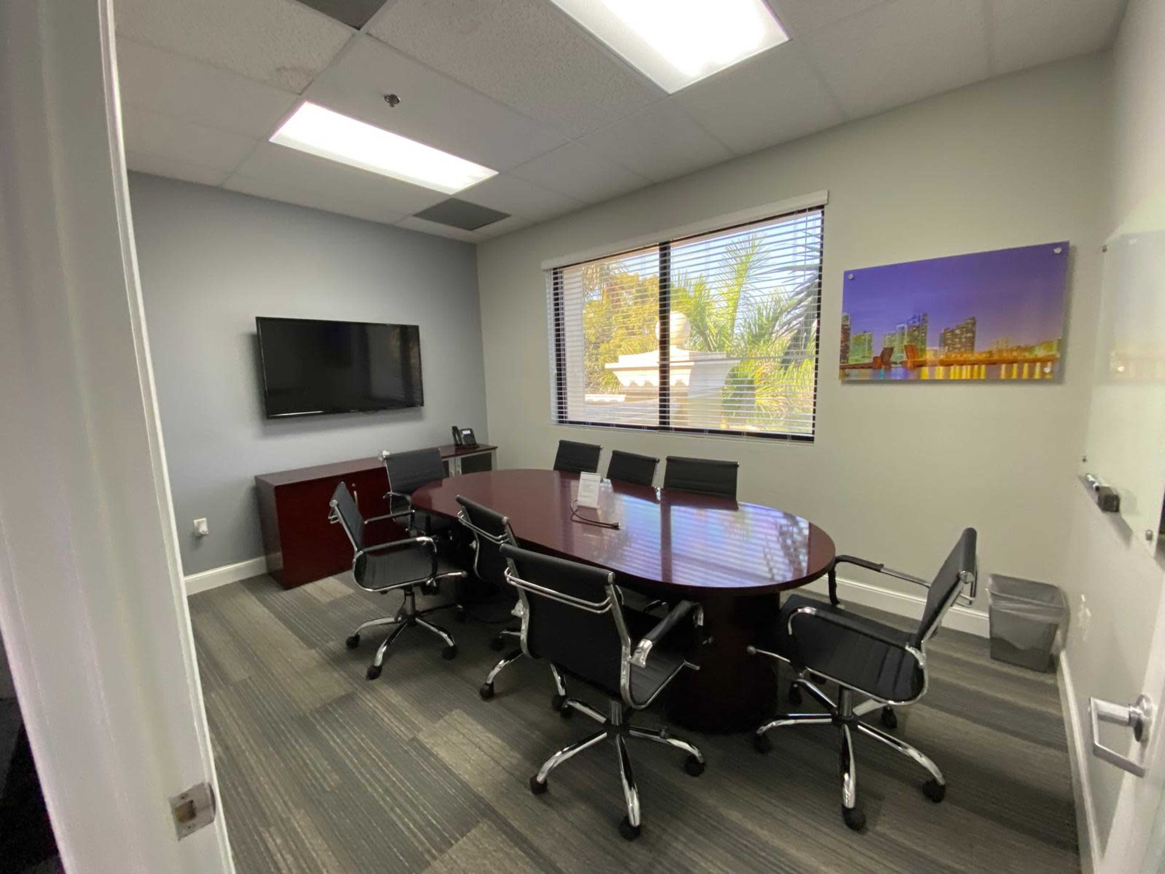 The image shows a conference room featuring a large oval table surrounded by six chairs, a wall-mounted television, and a window with blinds letting in natural light.
