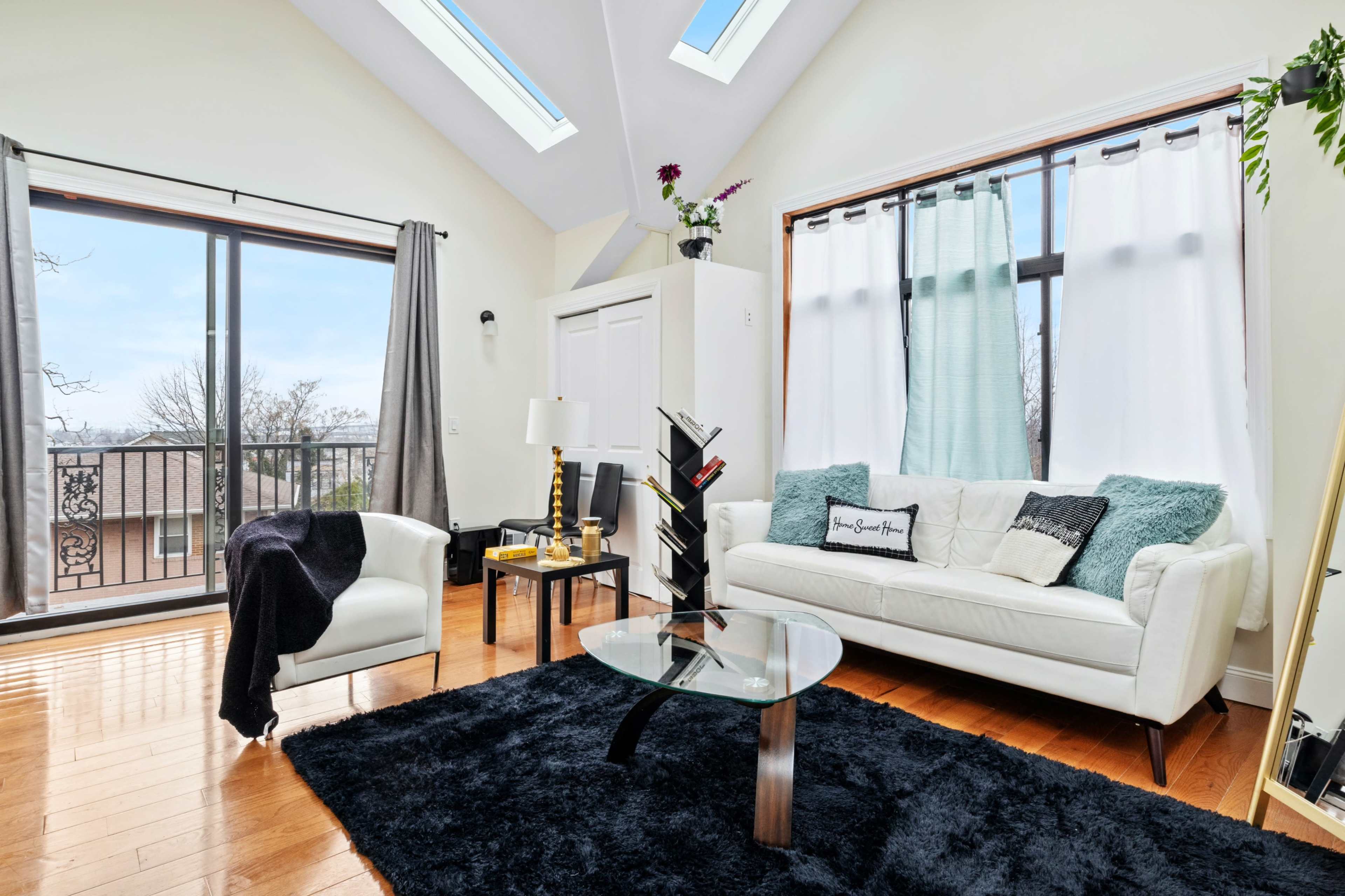 A bright living room features a white sofa, a black rug, large windows with views, and modern decor against a backdrop of wooden floors.
