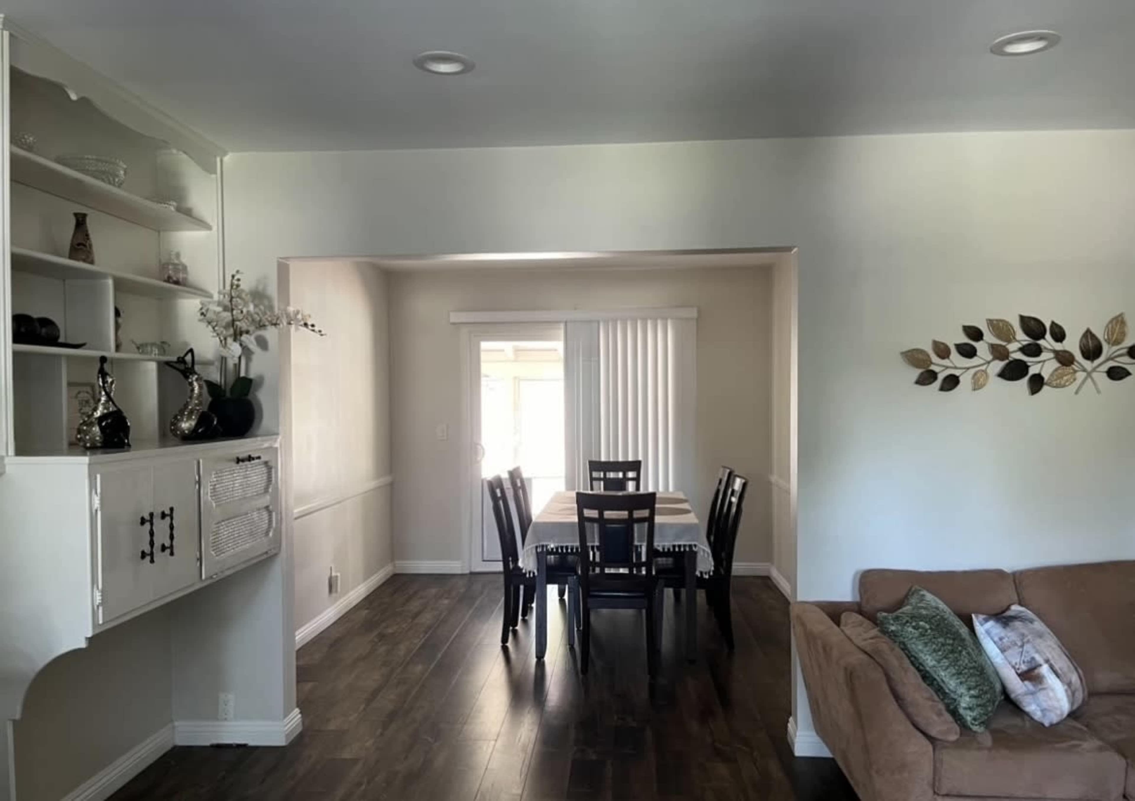 The image shows a bright, open living space with a dining area that features a wooden table and black chairs, adjacent to a cozy couch and shelves along the wall.