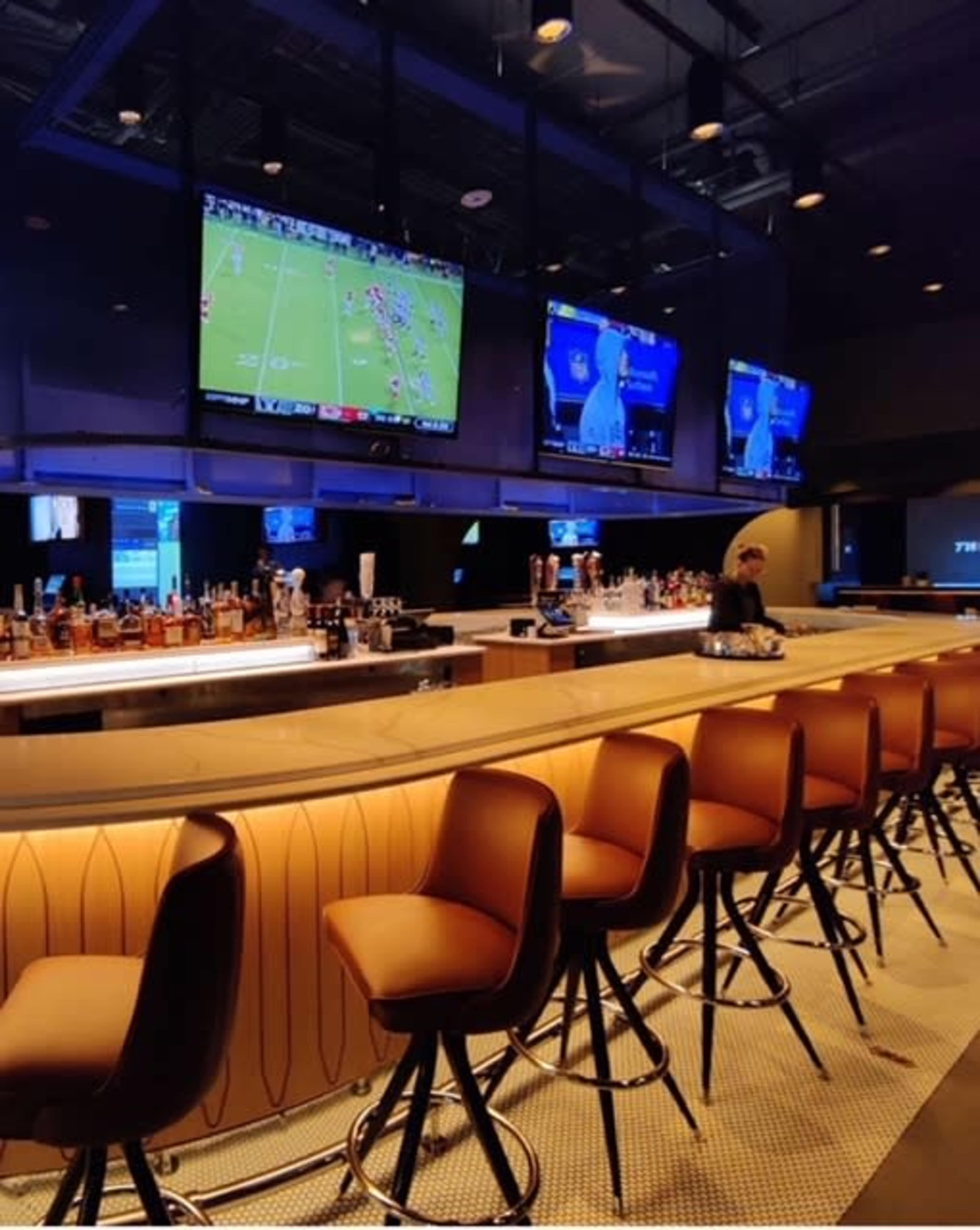 A modern bar with a long counter, several high stools, and multiple large screens displaying sports.