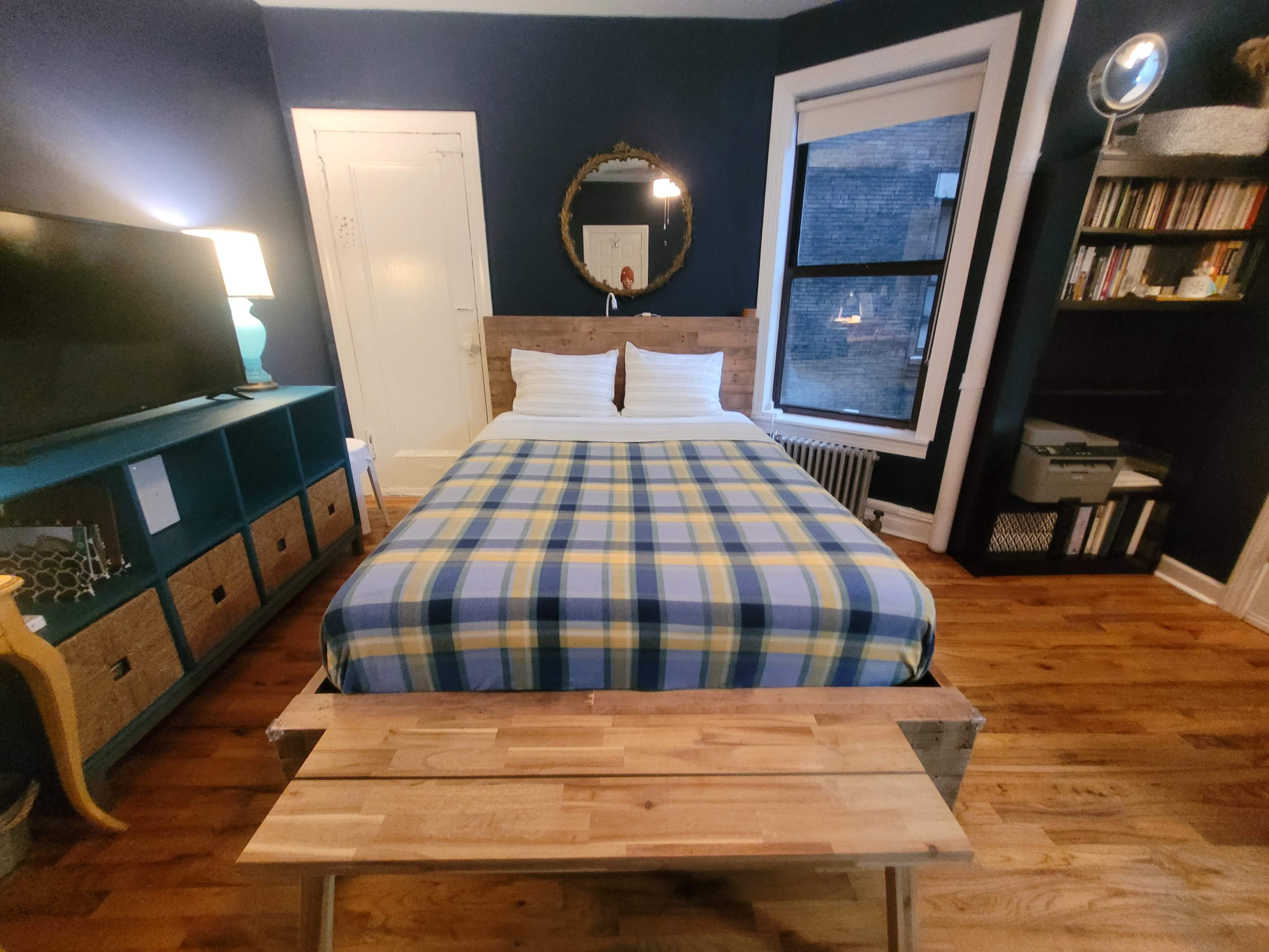 The image shows a compact bedroom featuring a bed with a plaid blanket, a television on a teal cabinet, and a window with natural light.