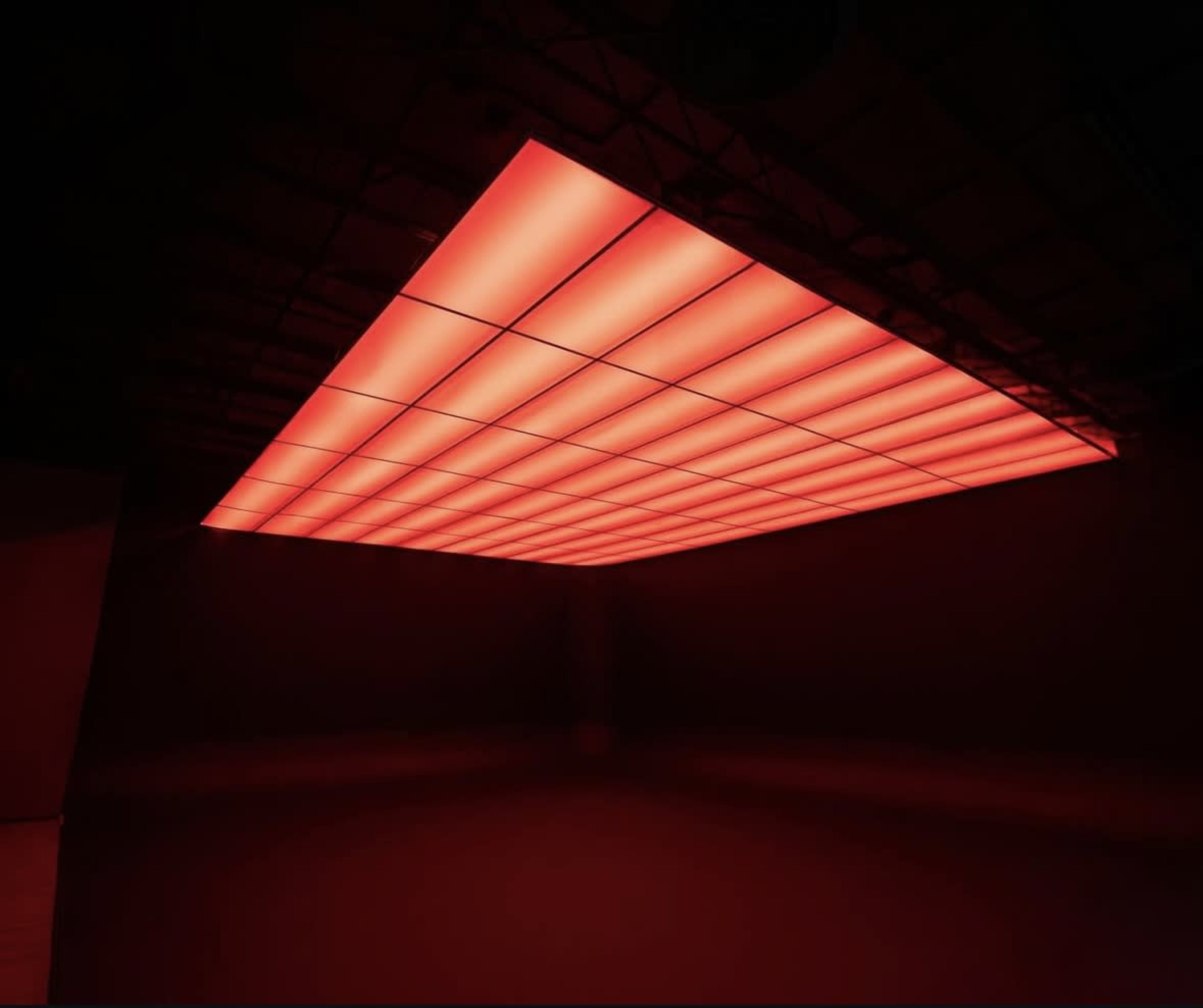 A large, rectangular ceiling panel comprised of grid-like sections emits a vibrant red light in a darkened space.