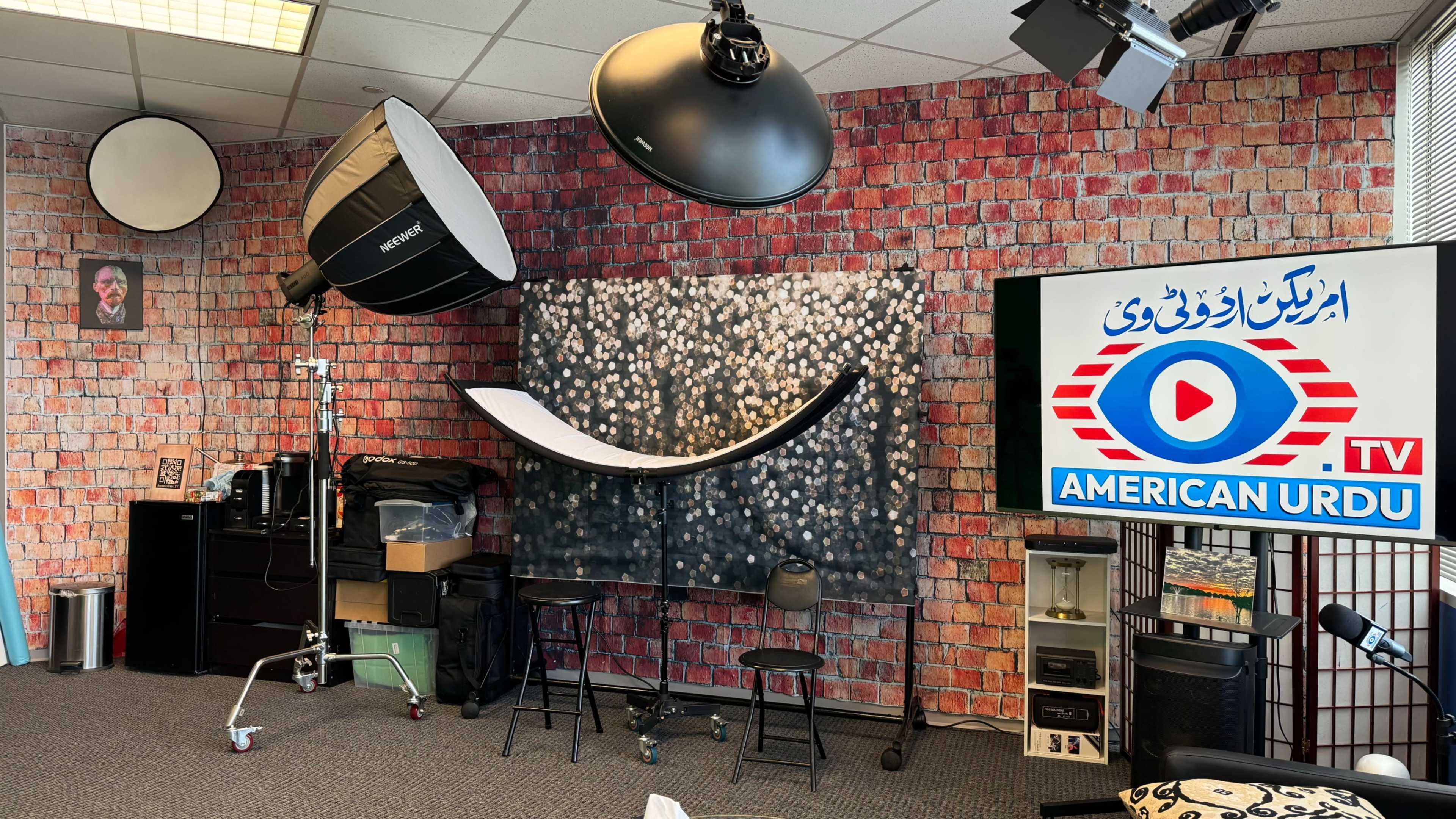 A studio setup with photography equipment, including softbox lights and a backdrop, alongside a television displaying a logo for "American Urdu TV."