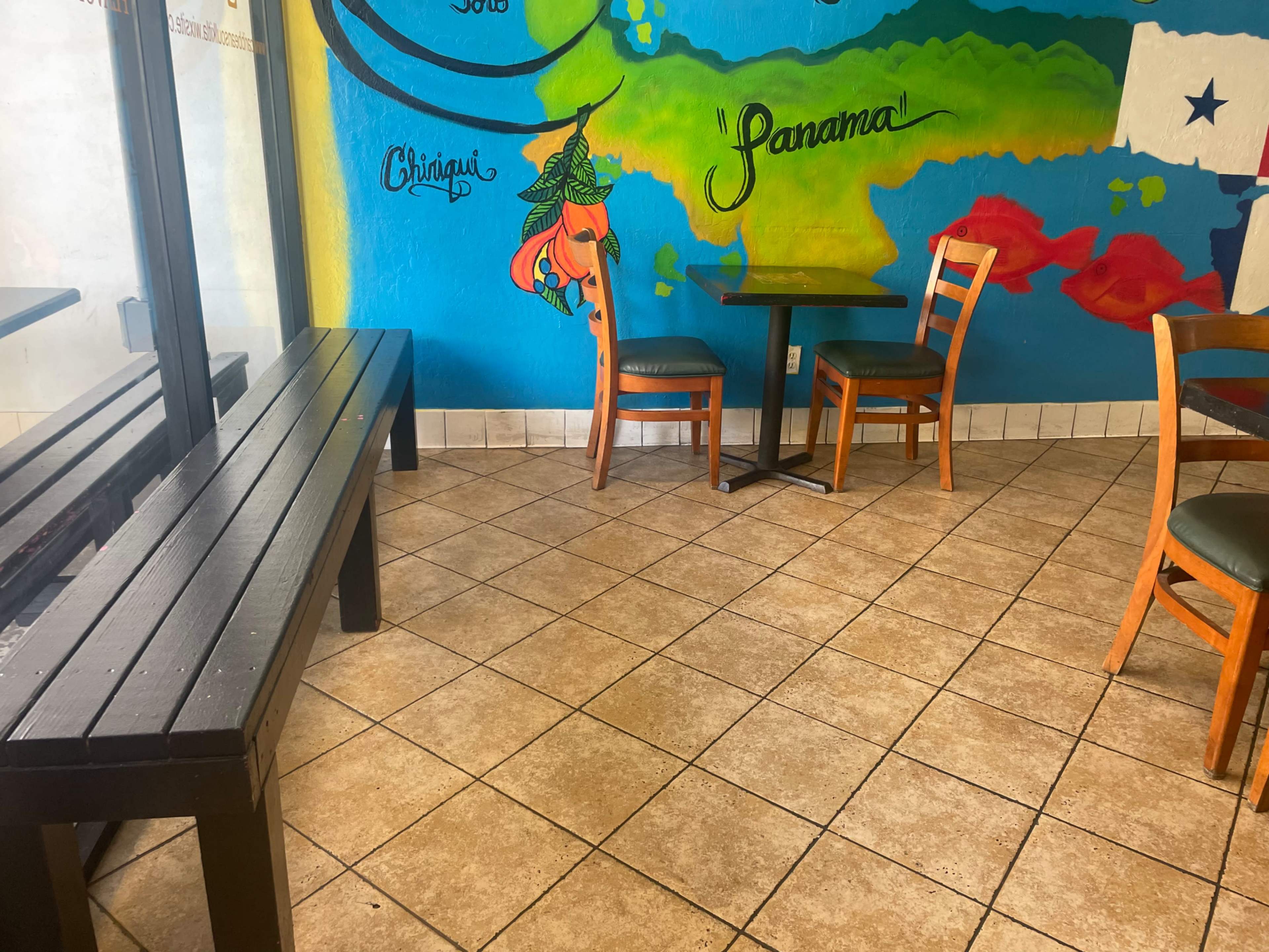 A small dining area with a bench, a table, and three wooden chairs against a colorful mural on the wall.