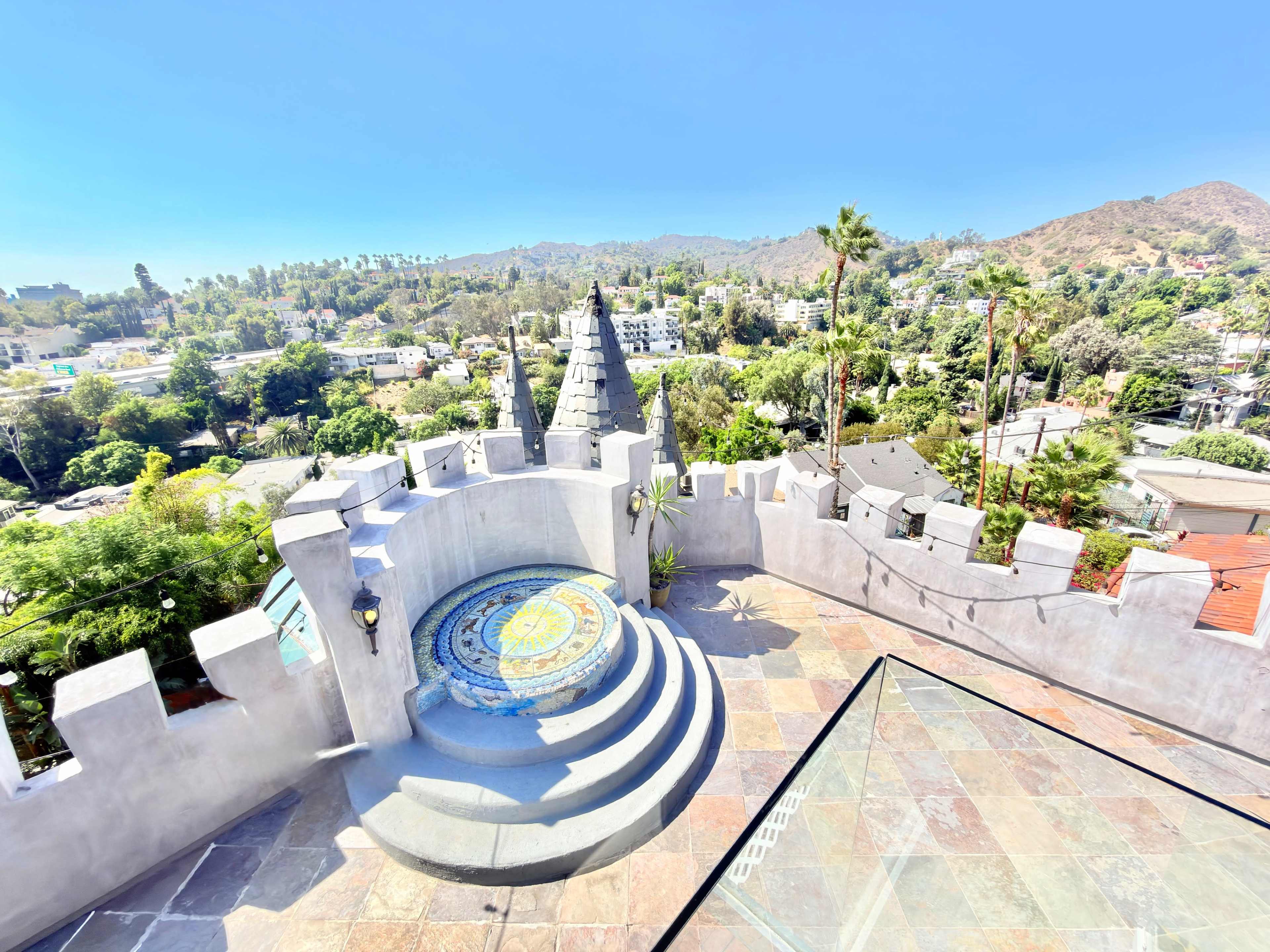 The image shows a stone terrace with a tiled circular seating area, surrounded by lush greenery and distant hills.