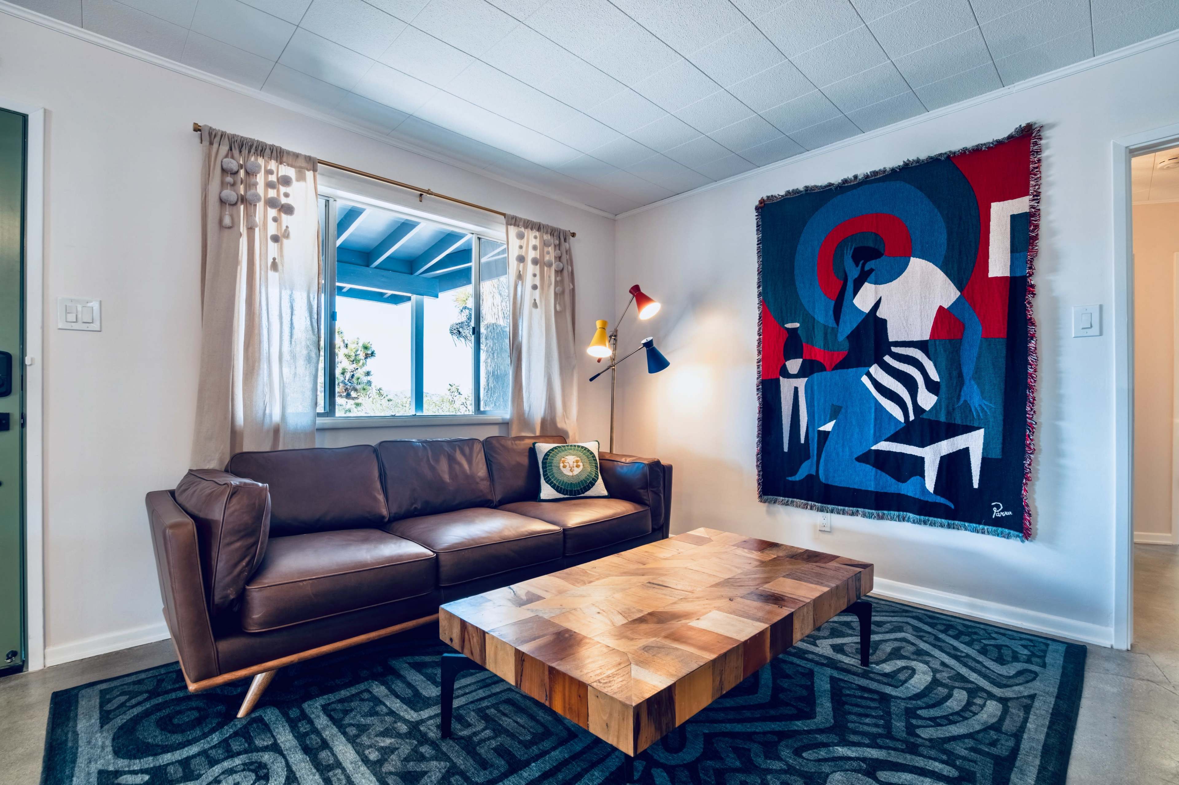 A leather couch sits across from a wooden coffee table, with a colorful artwork hanging on the wall beside a window adorned with curtains.