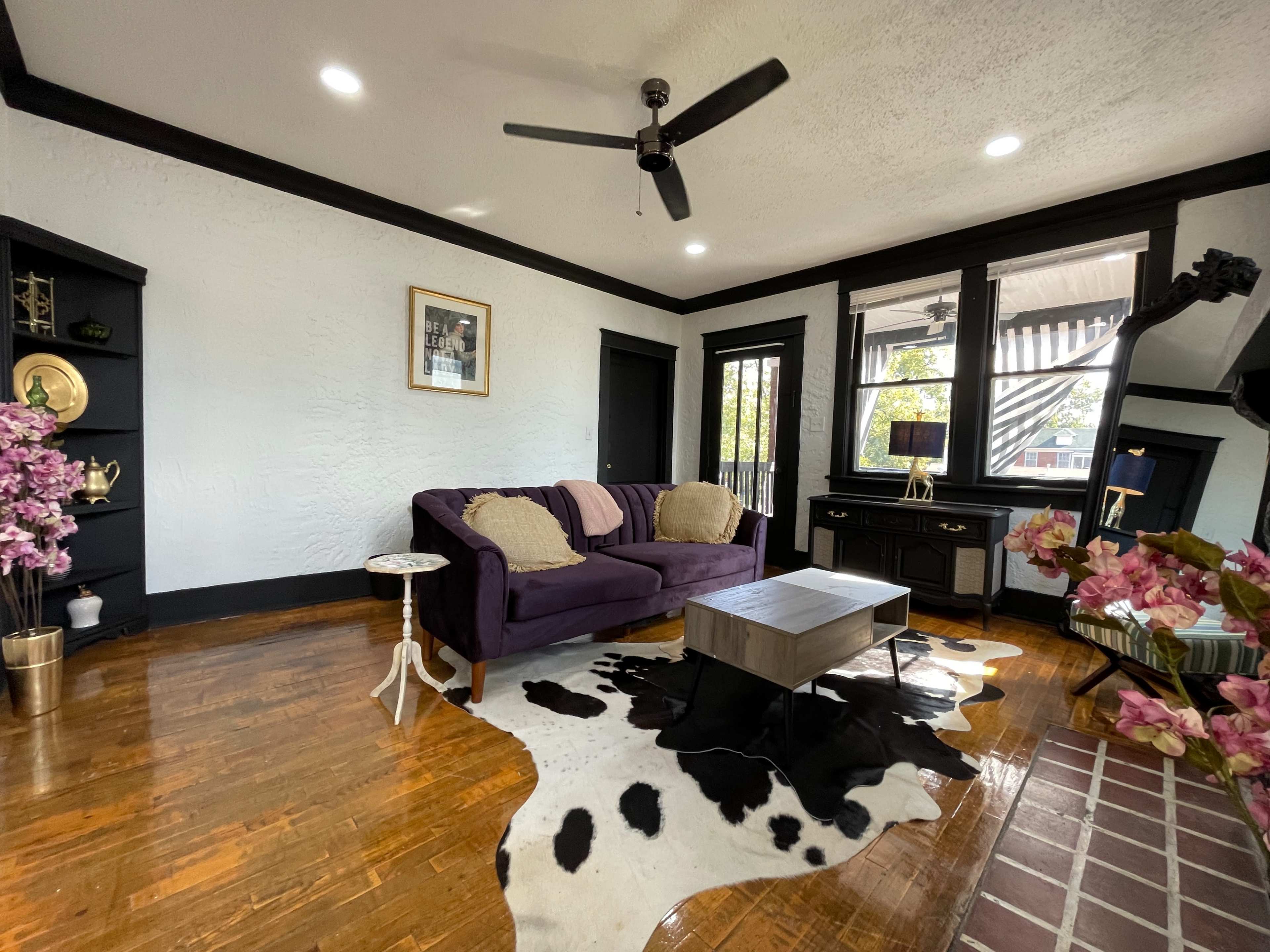 The living room features a purple sofa, a coffee table on a cowhide rug, and decorative elements against a backdrop of light walls and black trim.